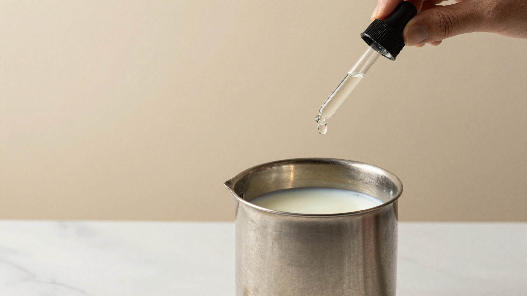 A glass dropper releasing a single drop of clear liquid enzyme into a stainless jug of milk