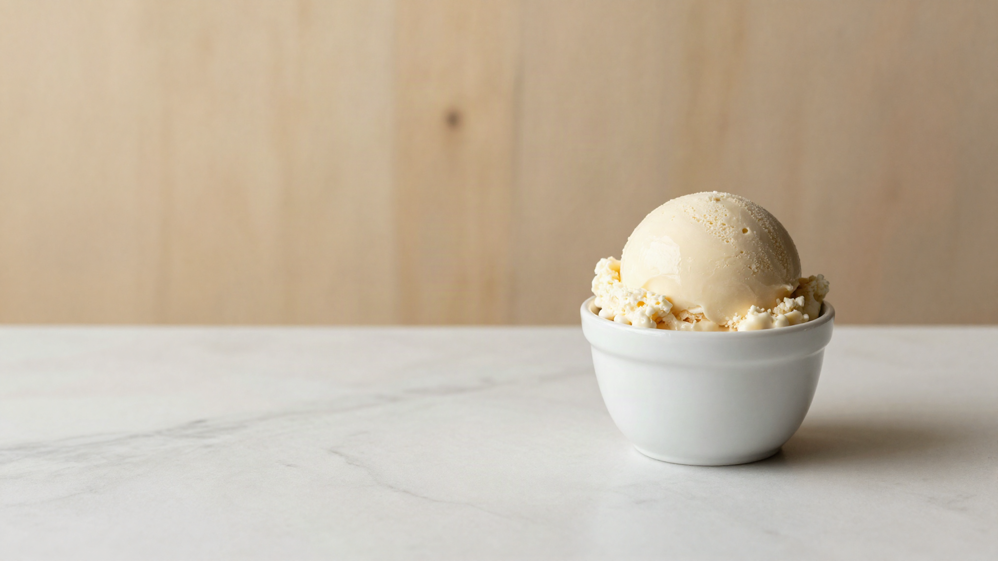 A single scoop of pale ivory gelato in a small white ceramic cup on marble