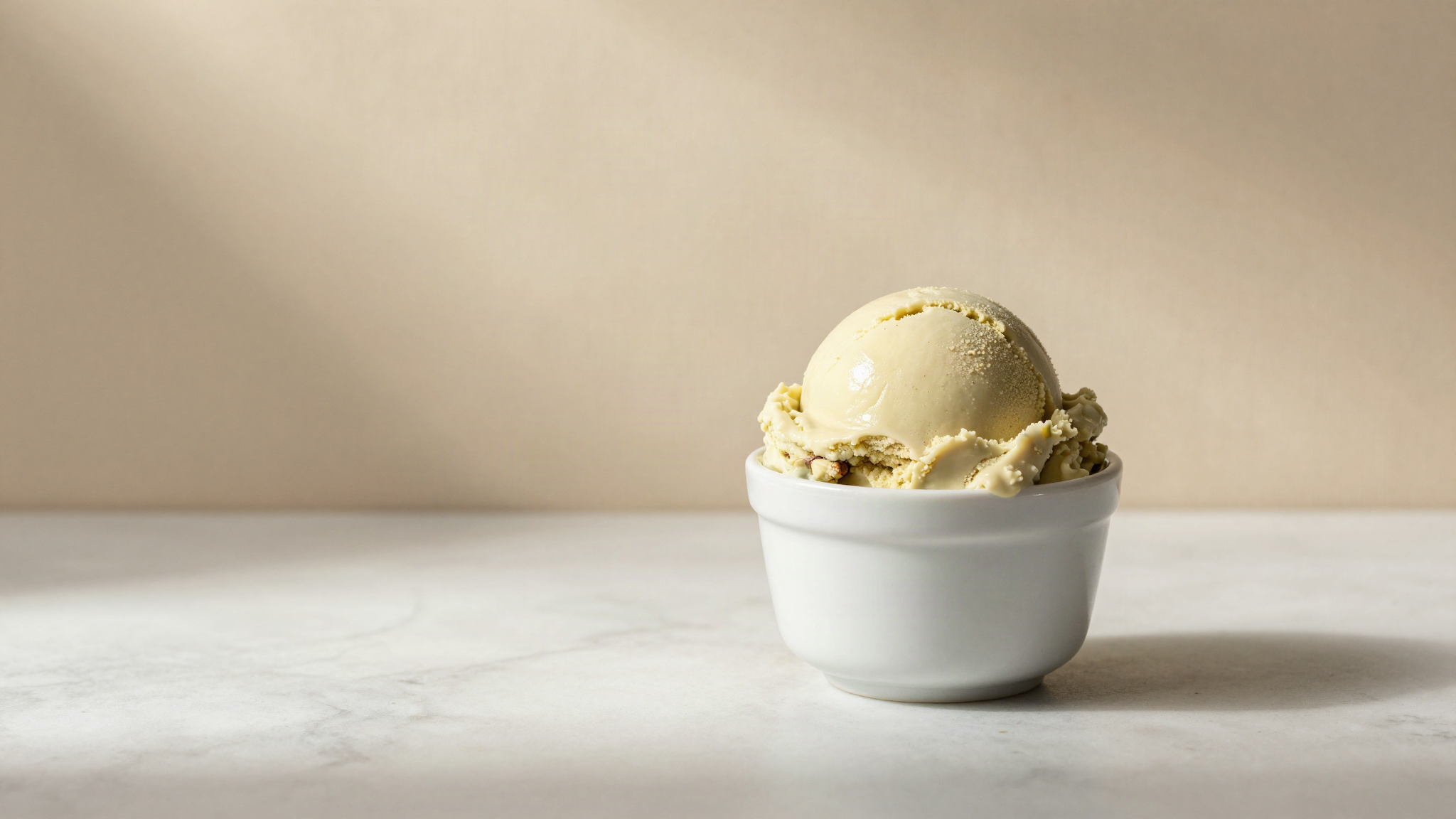 A dense glossy scoop of pale pistachio gelato in a small white ceramic cup on marble