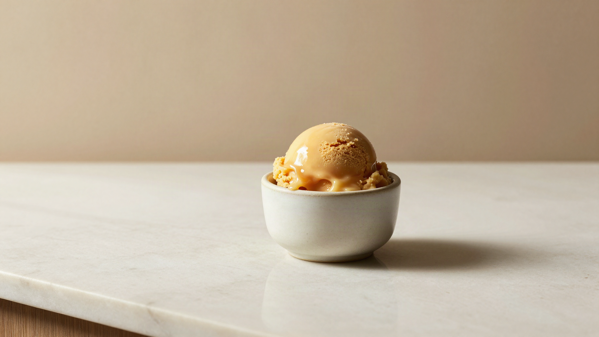 A dense glossy scoop of warm caramel toned maple gelato in a small ceramic cup on marble