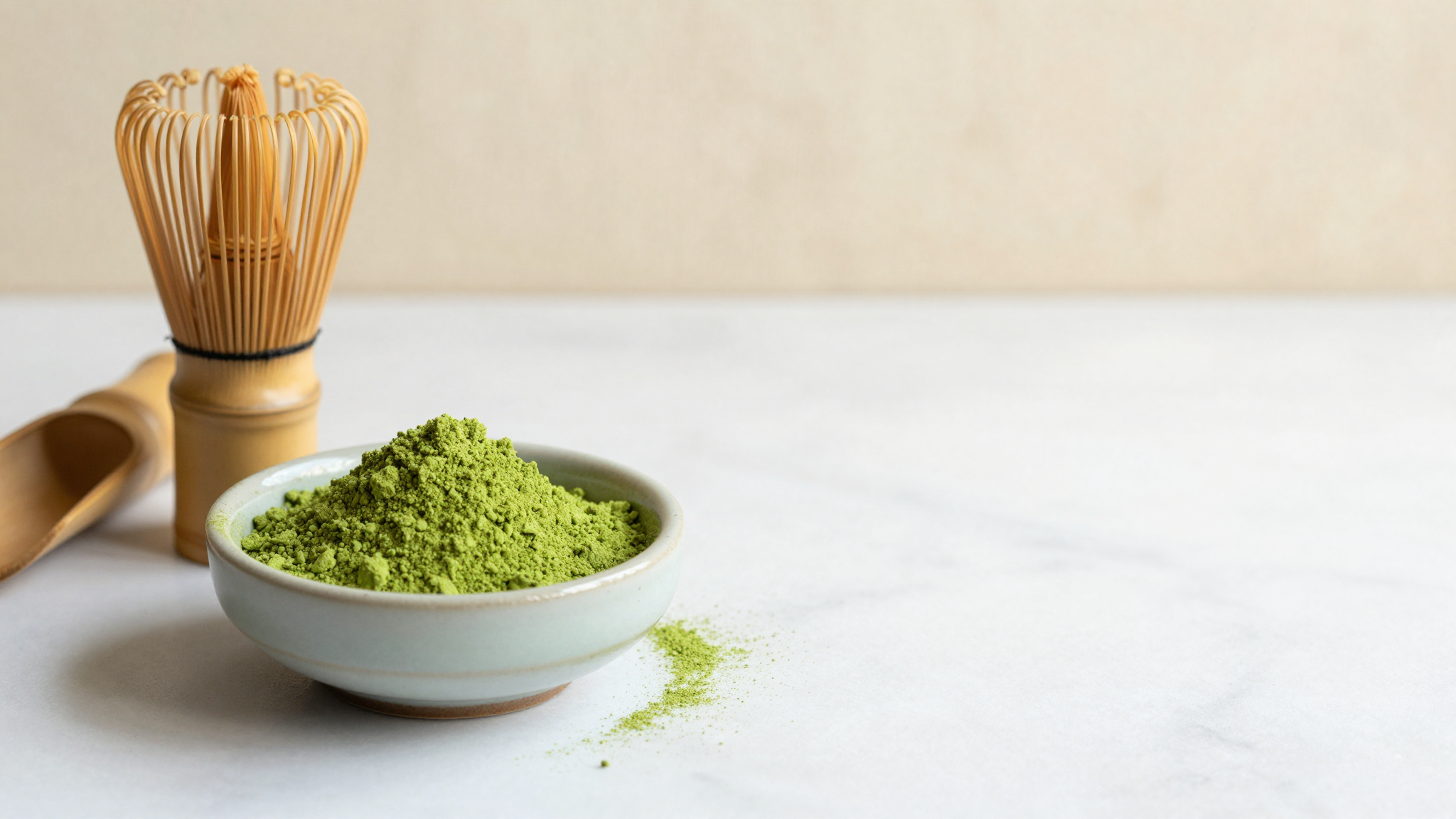 Vivid green matcha powder in a ceramic bowl beside a bamboo whisk and scoop on white marble
