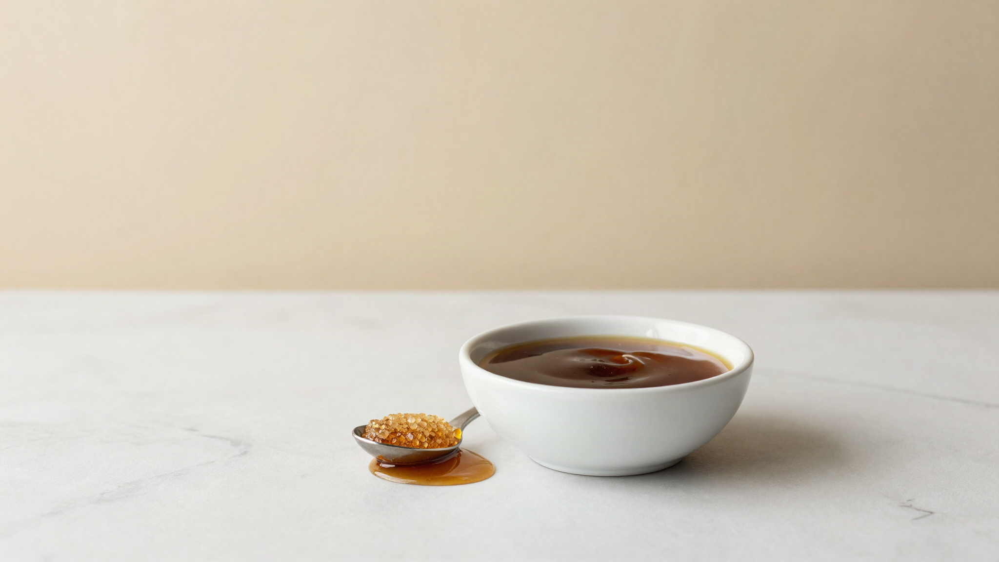 A dark glossy pool of molasses in a white ceramic bowl on marble beside a spoon of raw cane sugar