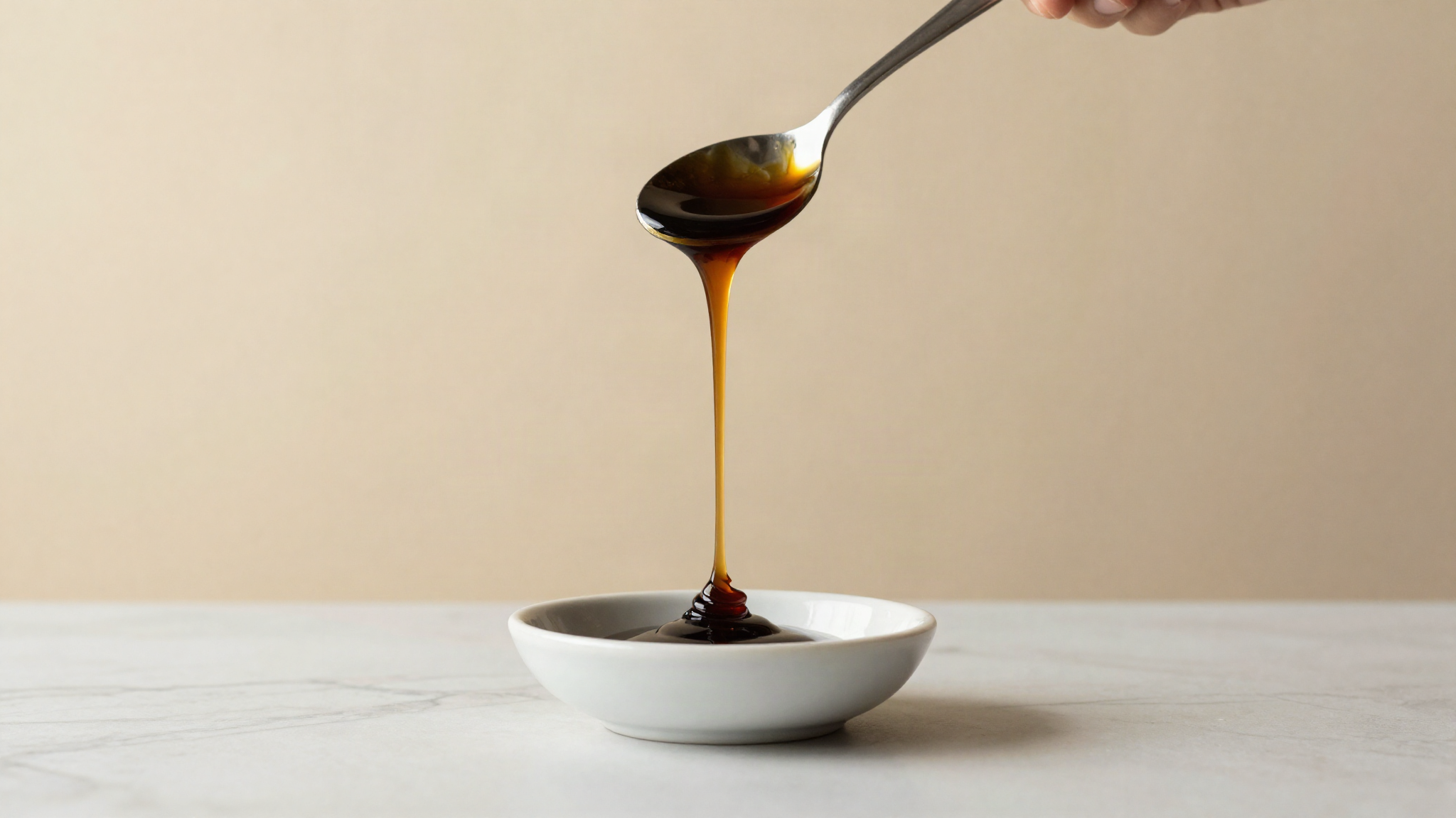 A dark ribbon of molasses falling slowly from a metal spoon into a small white bowl on a marble counter