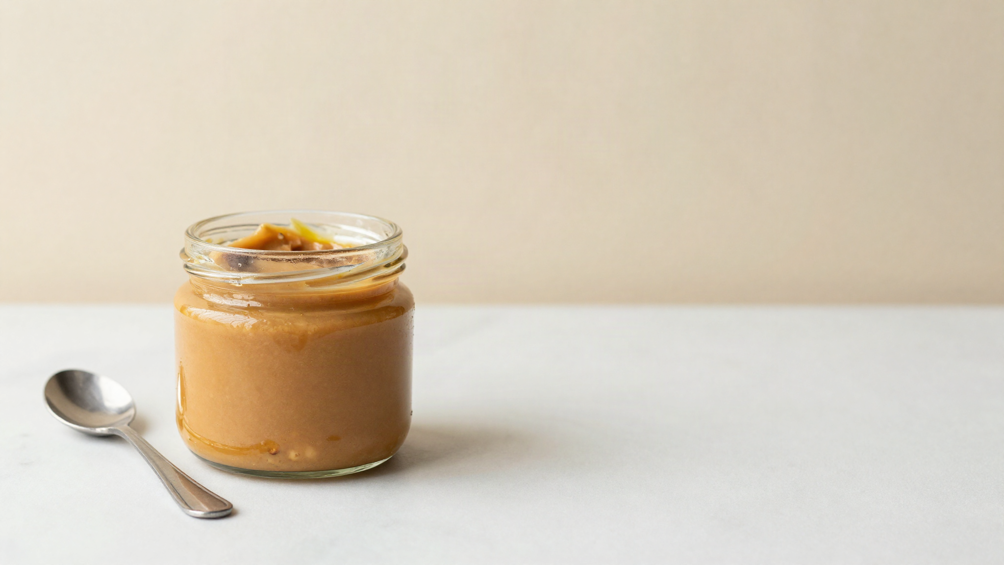 An open glass jar of smooth hazelnut paste with a thin golden layer of separated oil on top, on a marble counter beside a small spoon