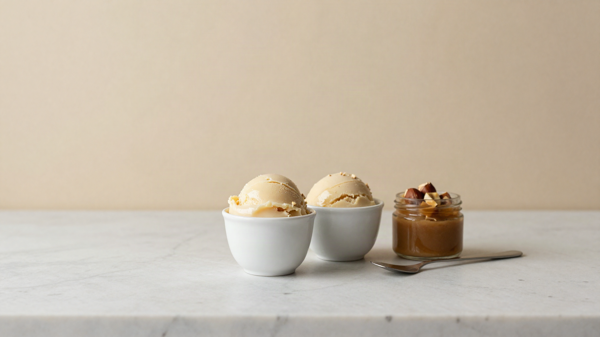 Two small white ceramic cups of hazelnut gelato on a marble counter, one slightly glossier than the other, beside an open jar of paste and a spatula