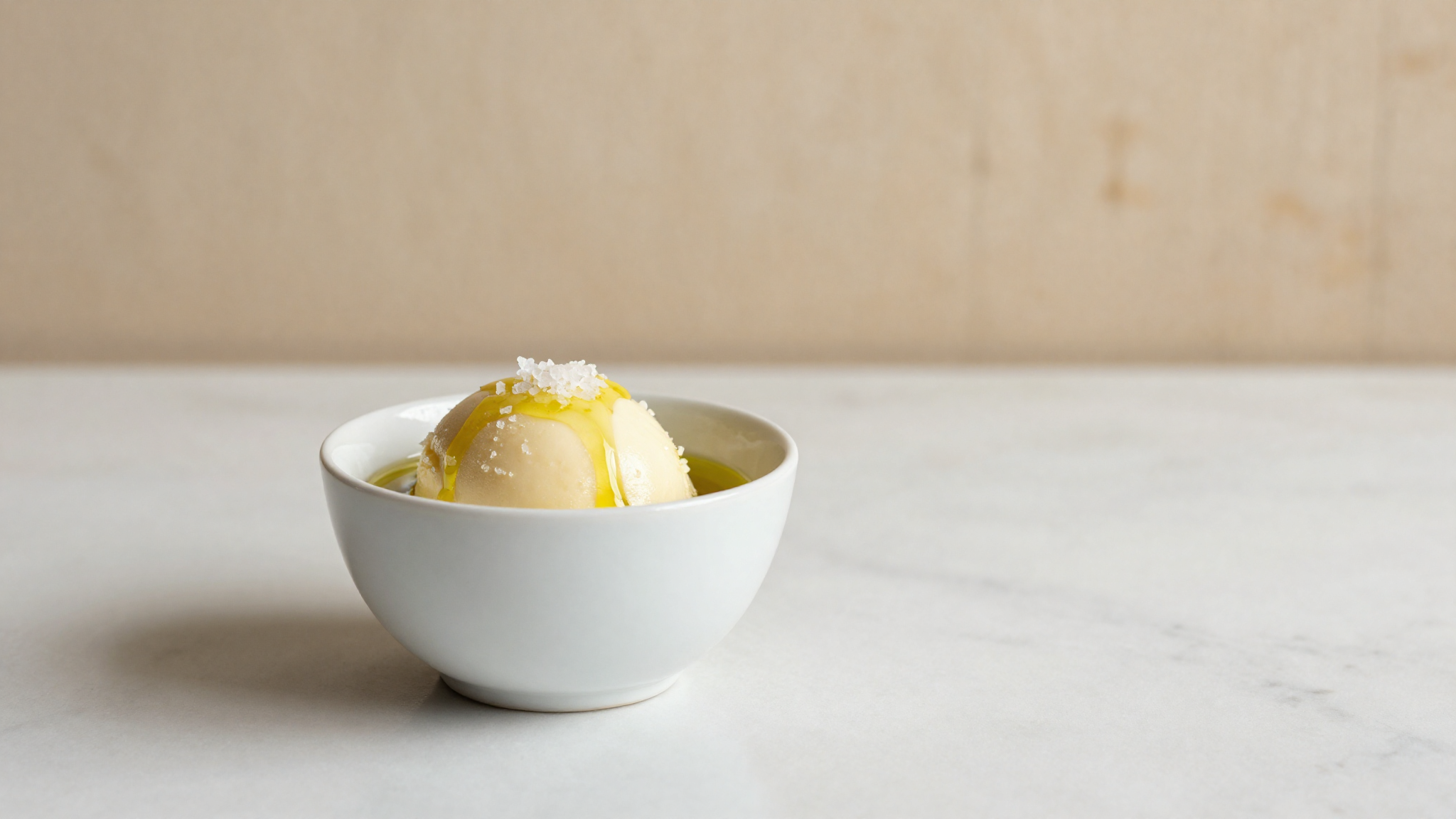 A finished serving of pale golden olive oil gelato drizzled with oil and Maldon salt flakes in a white ceramic cup