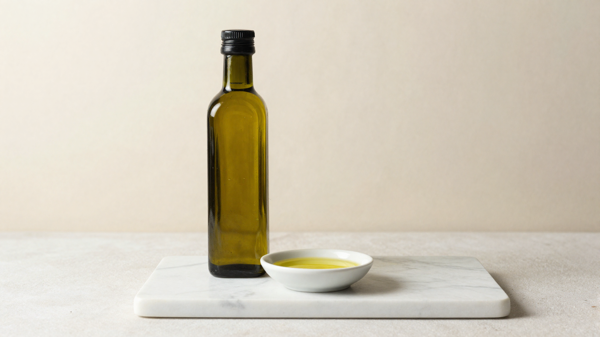 A bottle of extra virgin olive oil beside a small white ceramic dish of golden oil on white marble