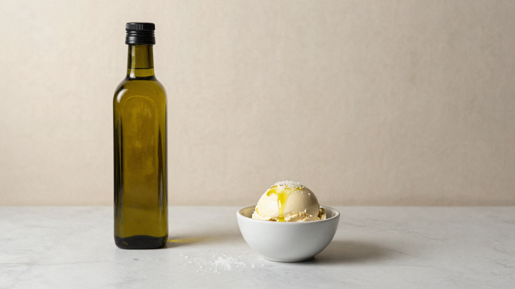 A bottle of extra virgin olive oil beside a scoop of pale gelato with sea salt on marble