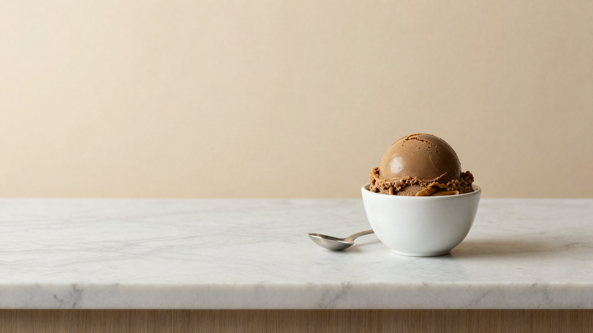 A scoop of deep caramel brown gelato in a small white ceramic cup with a silver spoon