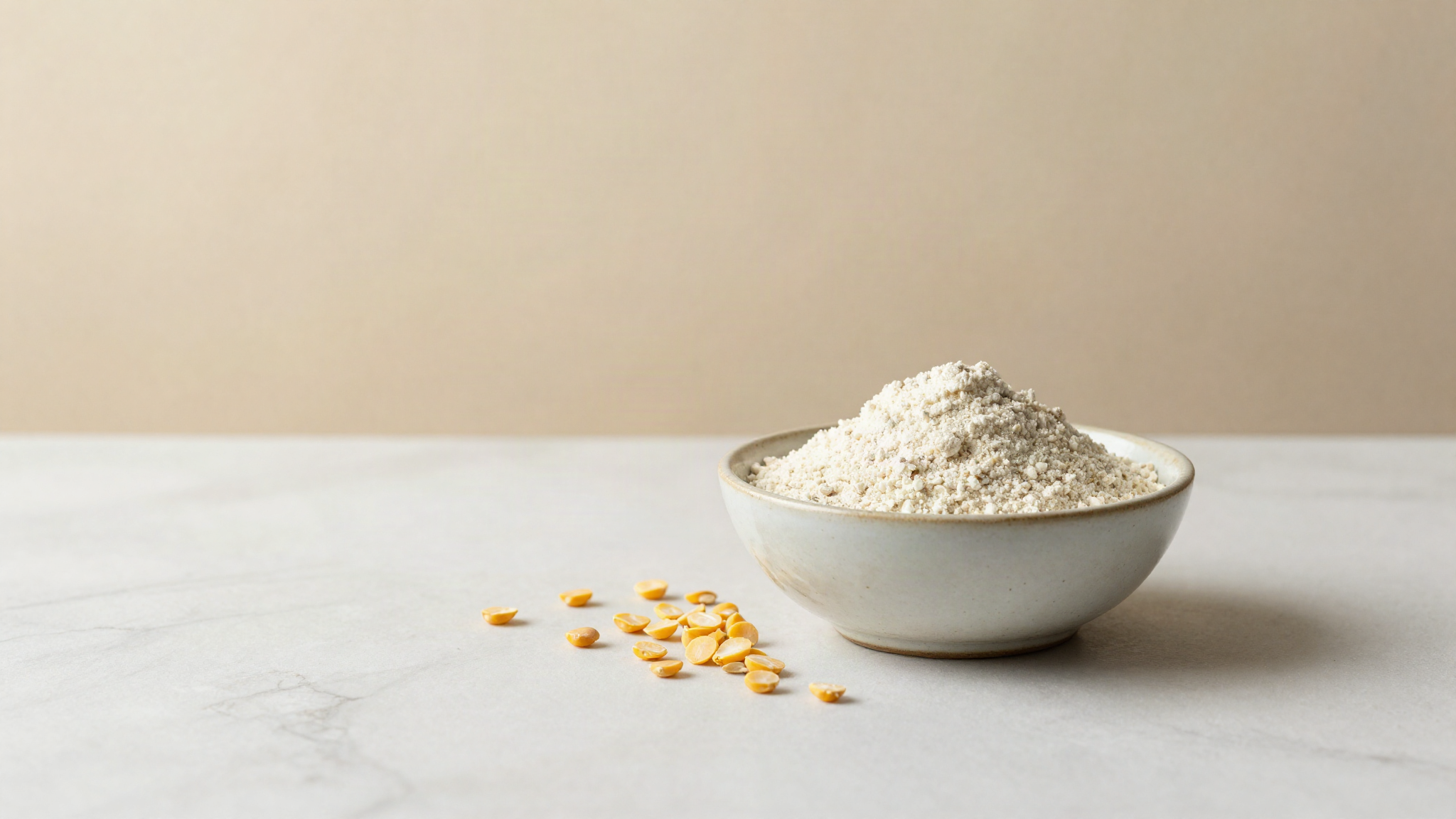 A small ceramic bowl of pale pea protein powder beside dried yellow split peas on a marble surface