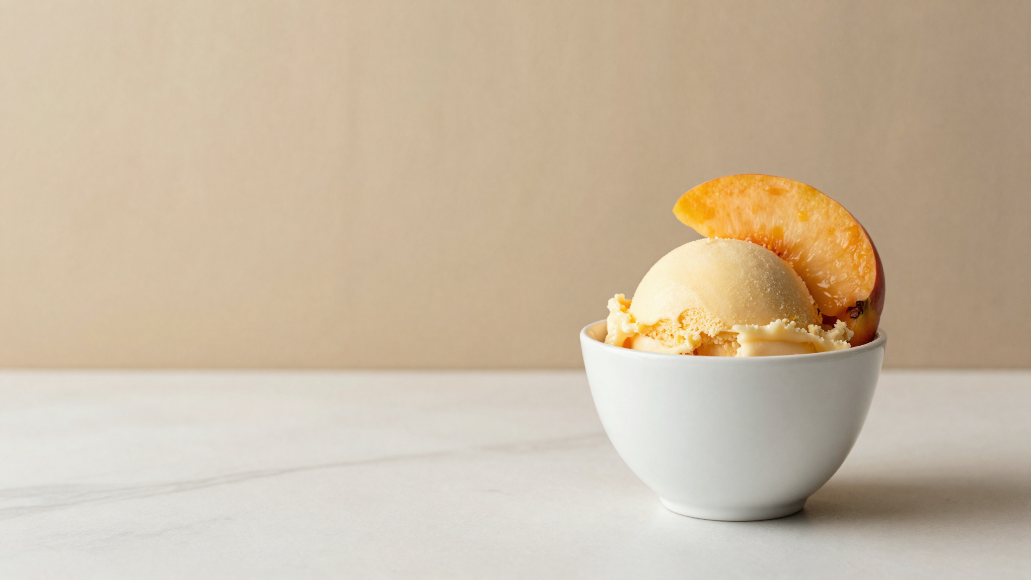 A serving of pale gold peach gelato in a white ceramic cup on a marble counter