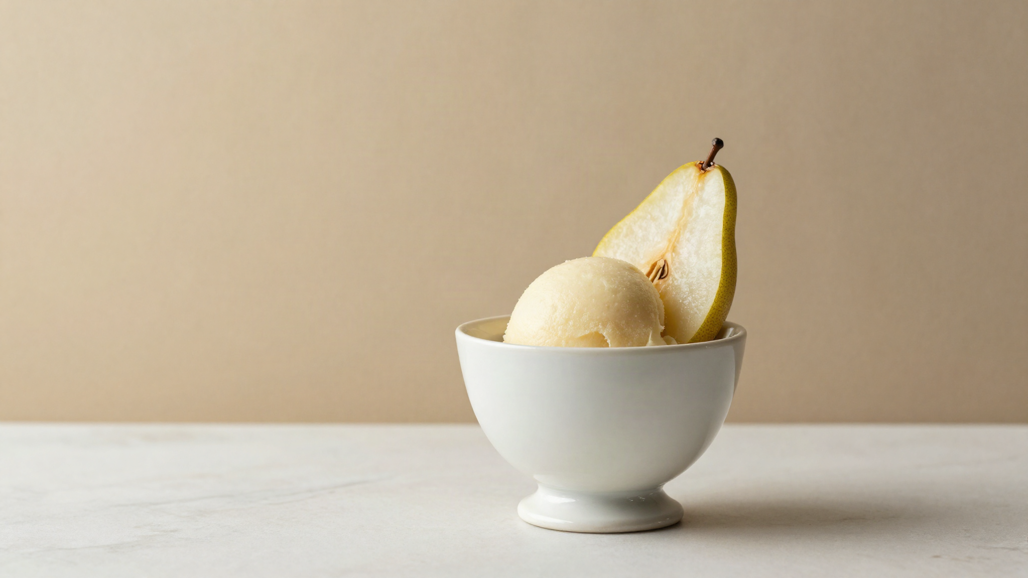 Pale ivory pear sorbetto served in a white ceramic cup with a thin fresh pear slice on marble