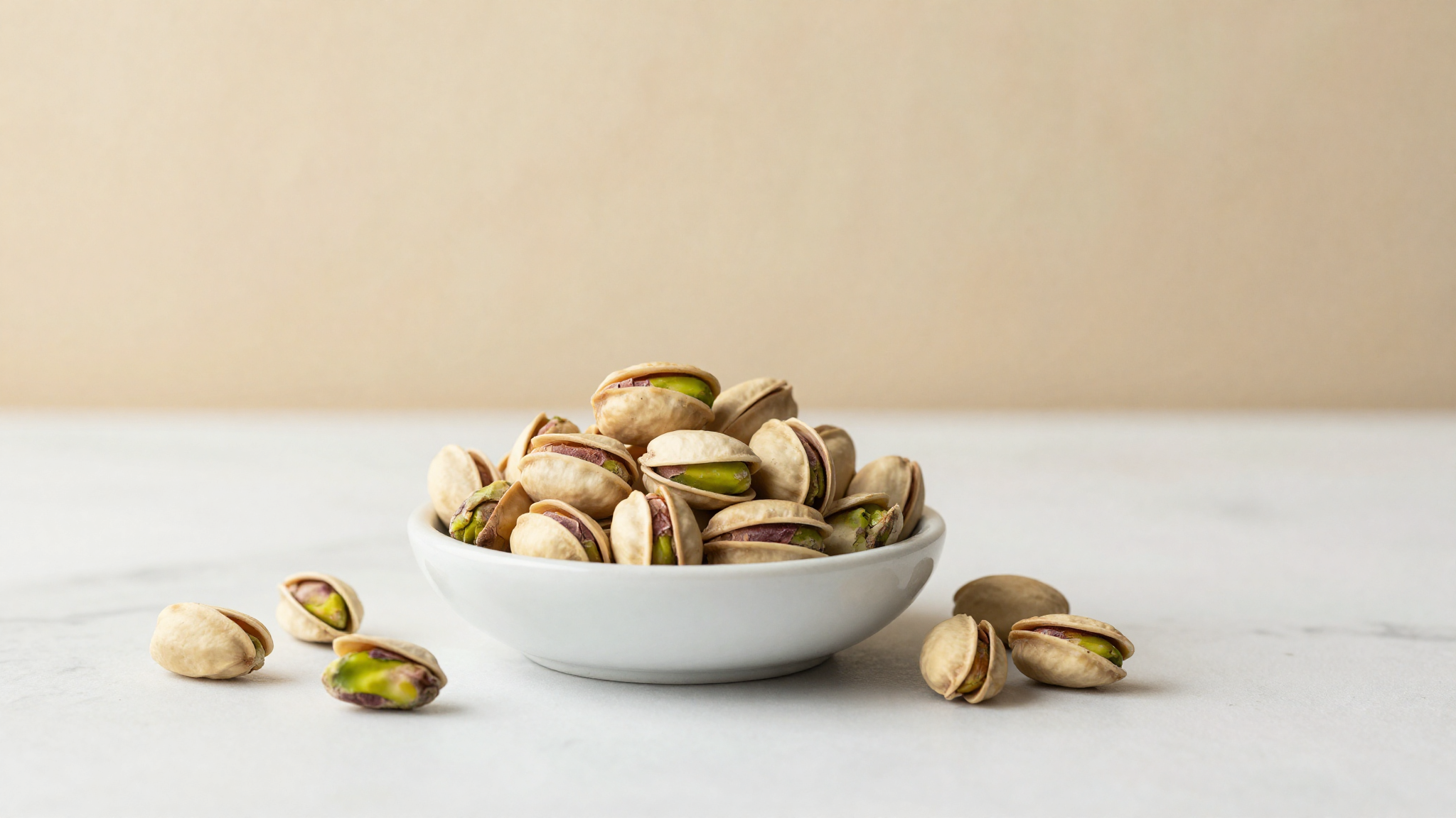 Fresh shelled green pistachios in a small white ceramic bowl on a marble surface with whole pistachios in shells nearby