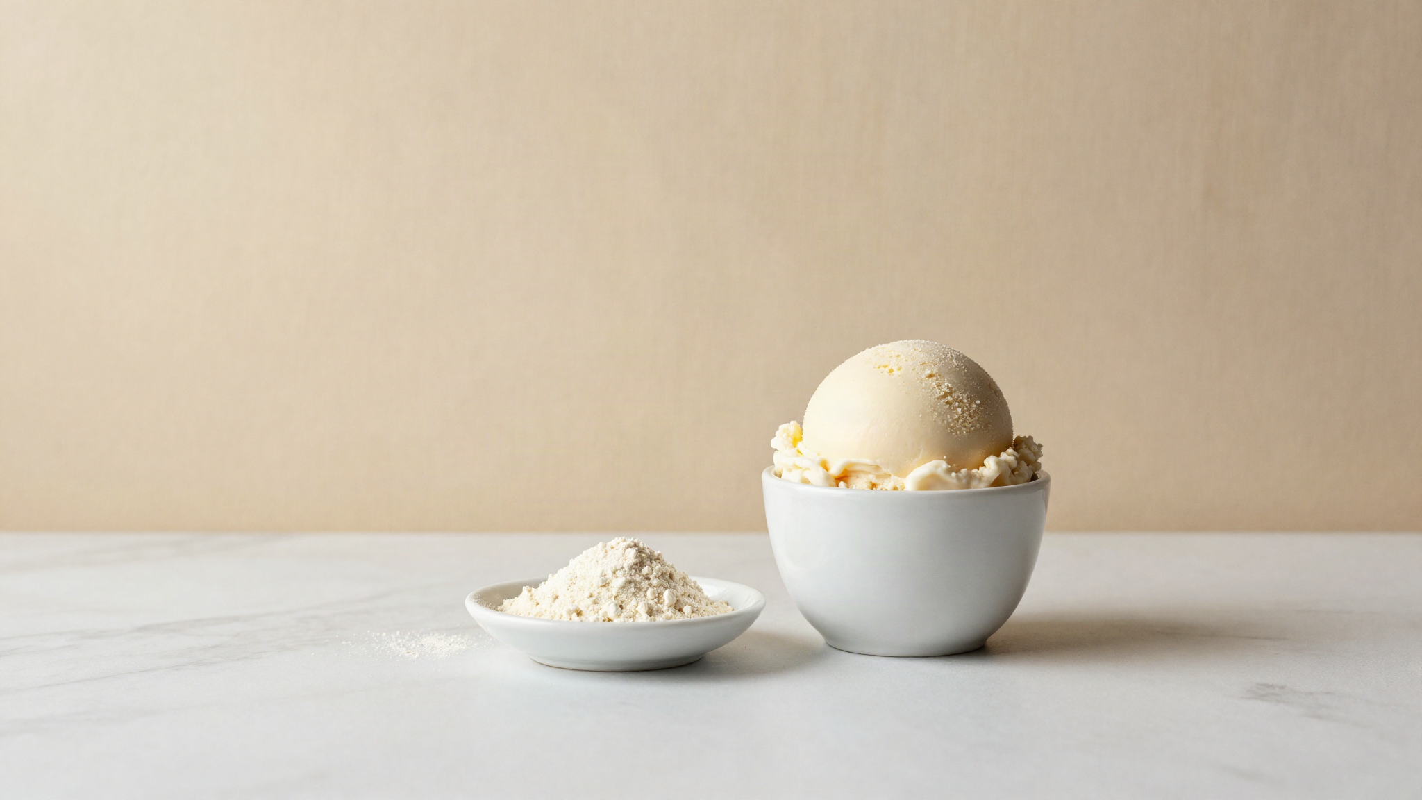 A single scoop of pale ivory gelato in a small white ceramic cup beside a small dish of off-white powder on marble