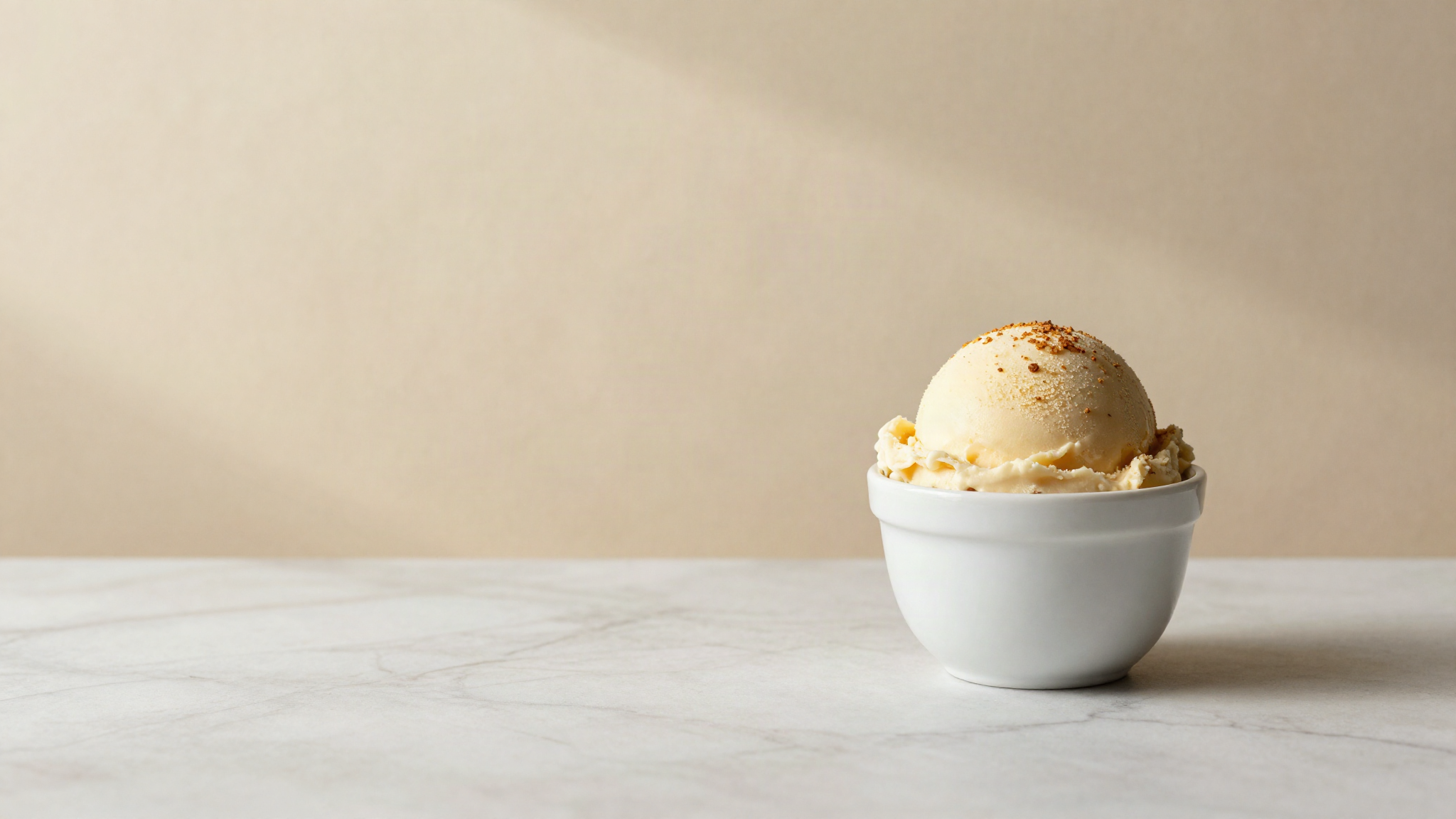 A scoop of pale italian gelato in a small white ceramic cup on marble with a faint golden cast