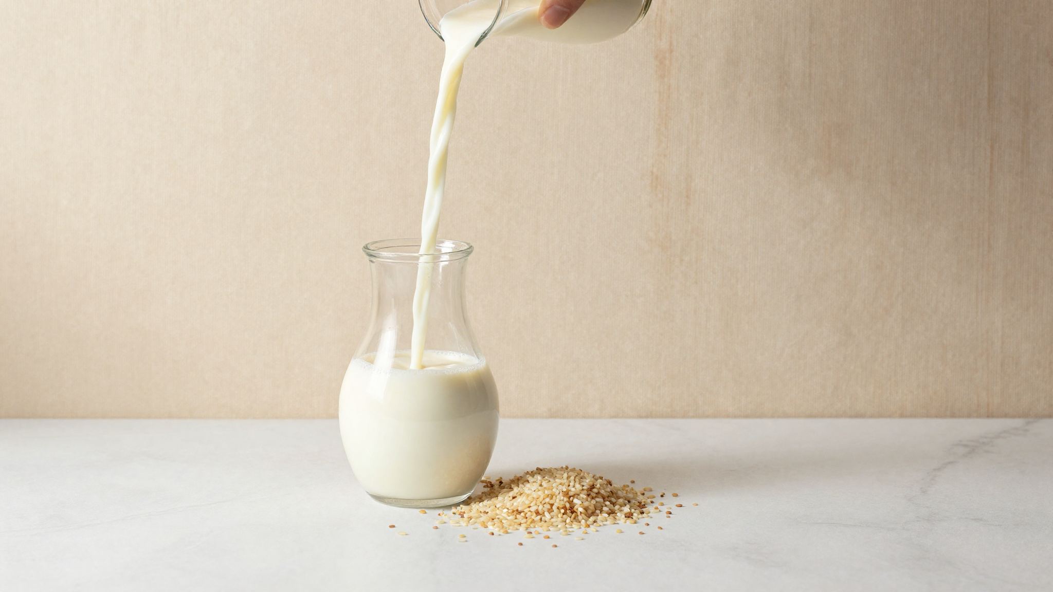 Rice milk being poured into a glass jug beside scattered raw rice grains on marble