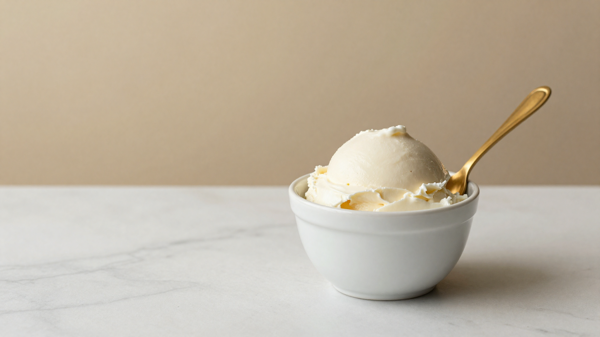 A smooth pale ricotta gelato in a white ceramic cup with a spoon on marble