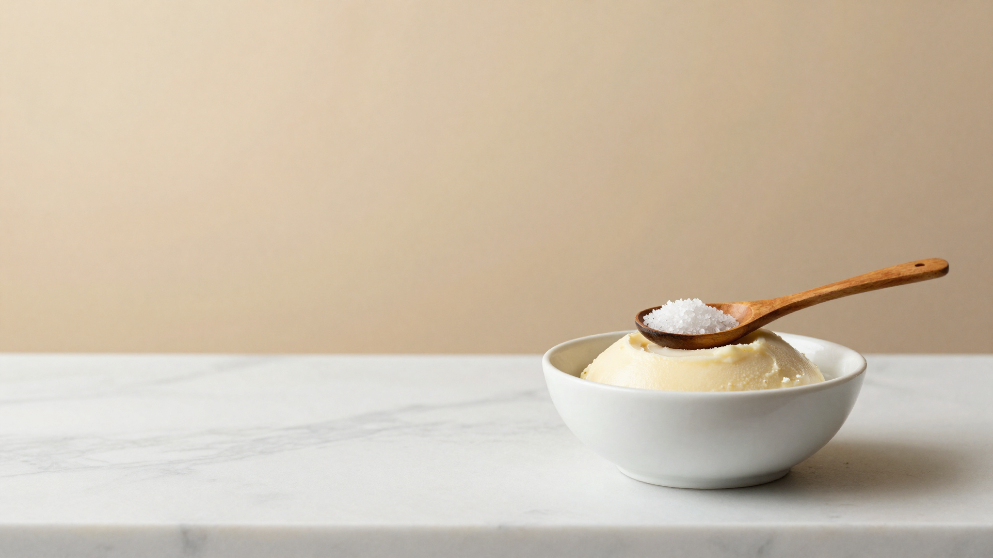 Fine sea salt in an olive-wood spoon held over a bowl of pale gelato base on marble
