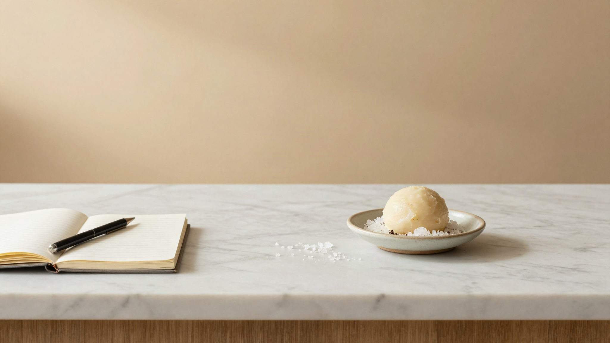 Marble counter seen from above with a notebook, a small precision scale, and ceramic dishes of flaky salt beside a pale savory gelato quenelle