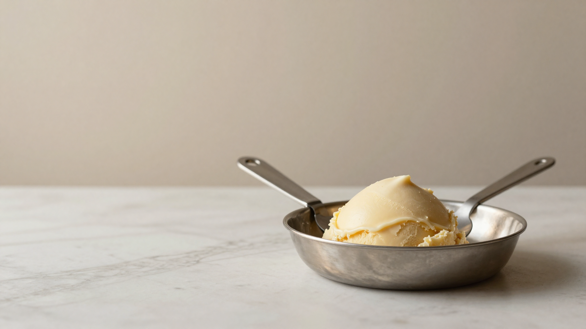 A stainless steel gelato spade and a portion disher resting on a marble counter beside a gelato pan