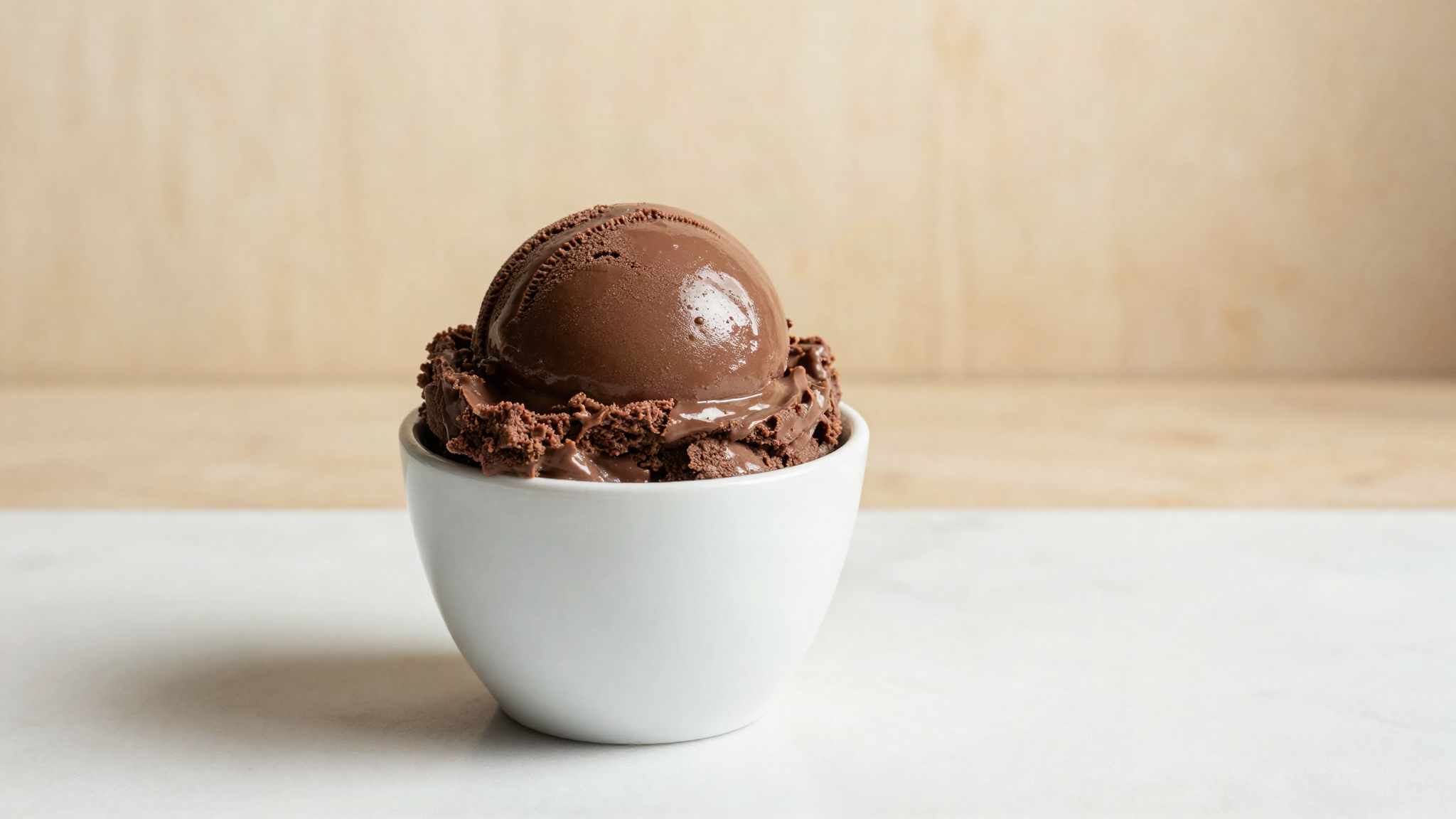 A glossy smooth scoop of dark chocolate gelato in a small white ceramic cup on a marble surface