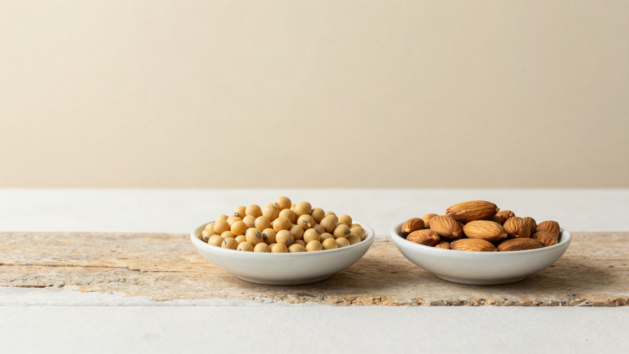 A small dish of dry soybeans and a small dish of whole raw almonds side by side on aged wood