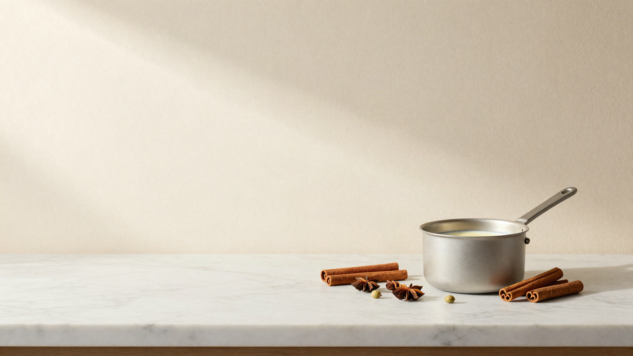 Whole spices arranged on a marble counter beside a small saucepan of warm milk