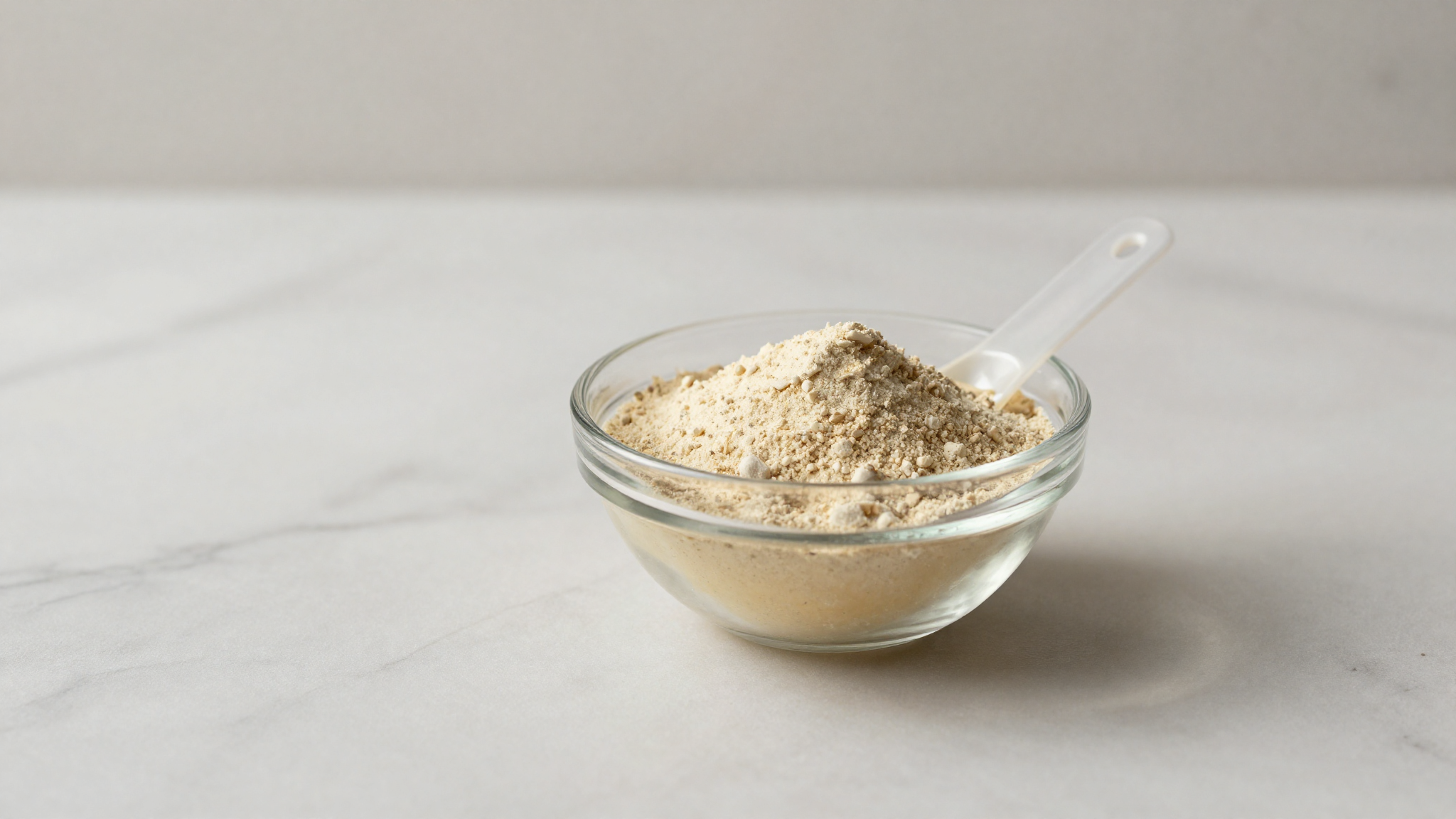 A small glass bowl of pale locust bean gum powder with a measuring spoon on marble
