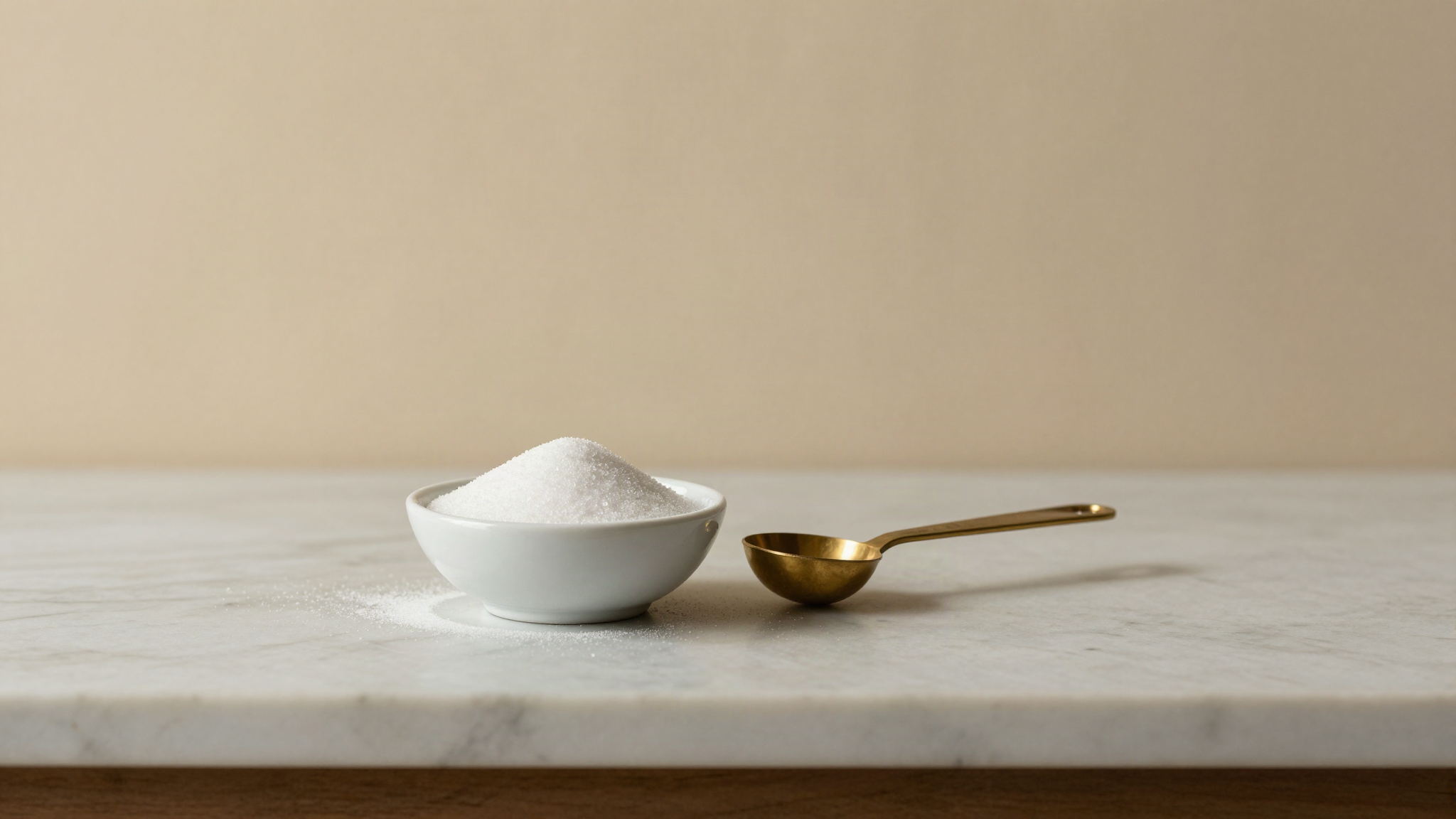 A small white ceramic bowl of sugar dry-blended with stabilizer powder, with a brass measuring spoon beside it on marble