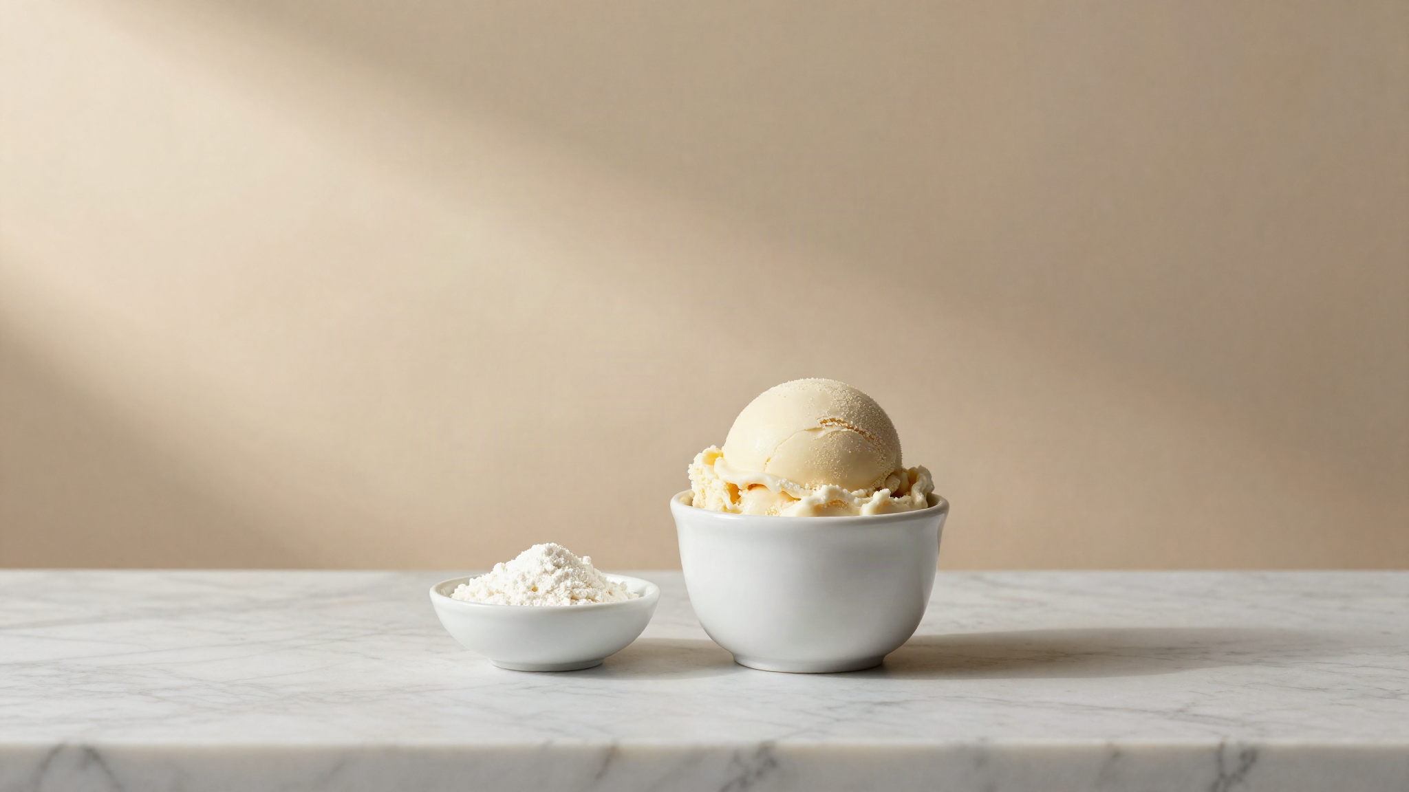 A finished scoop of smooth pale gelato in a white ceramic cup on a marble counter beside a small bowl of cornstarch