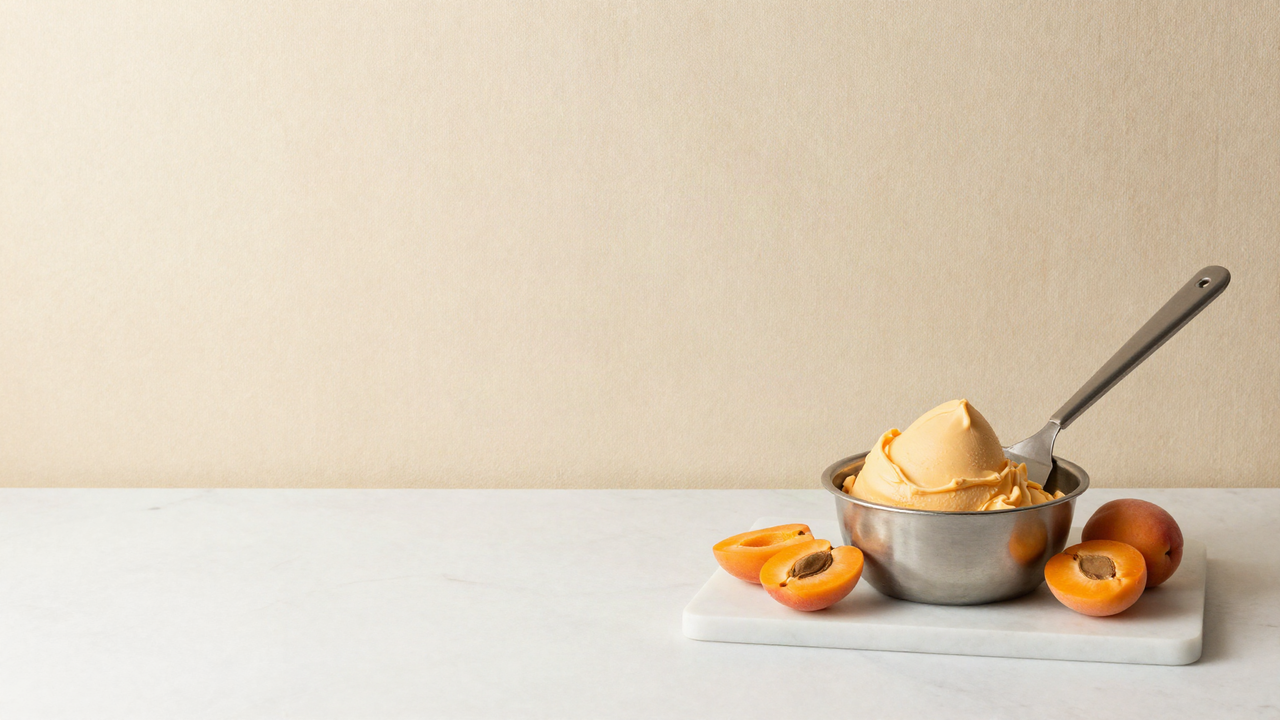 A stainless steel pan of freshly churned apricot gelato with a flat steel spatula and halved apricots beside it on marble