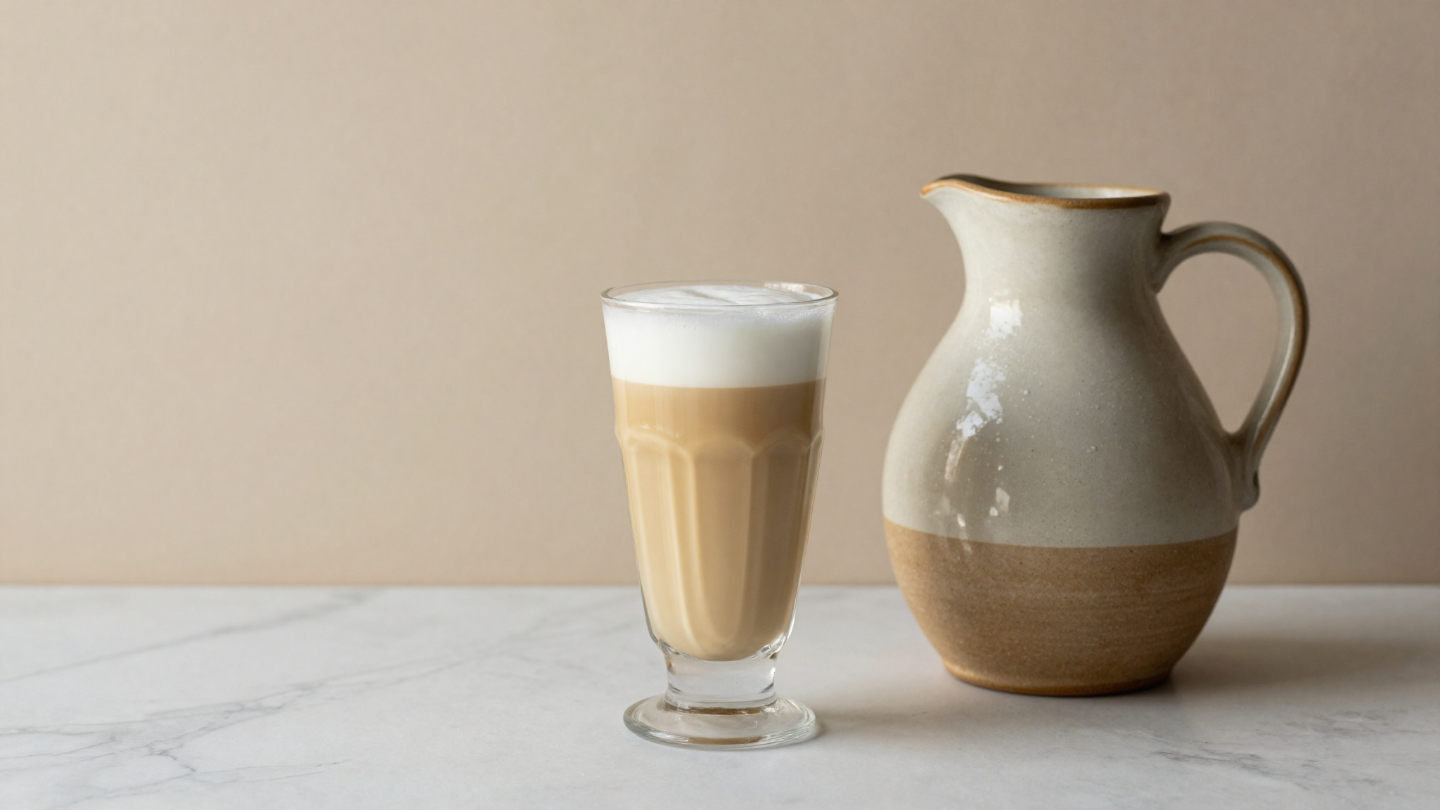 A glass of cultured Italian buttermilk beside a ceramic jug on white marble