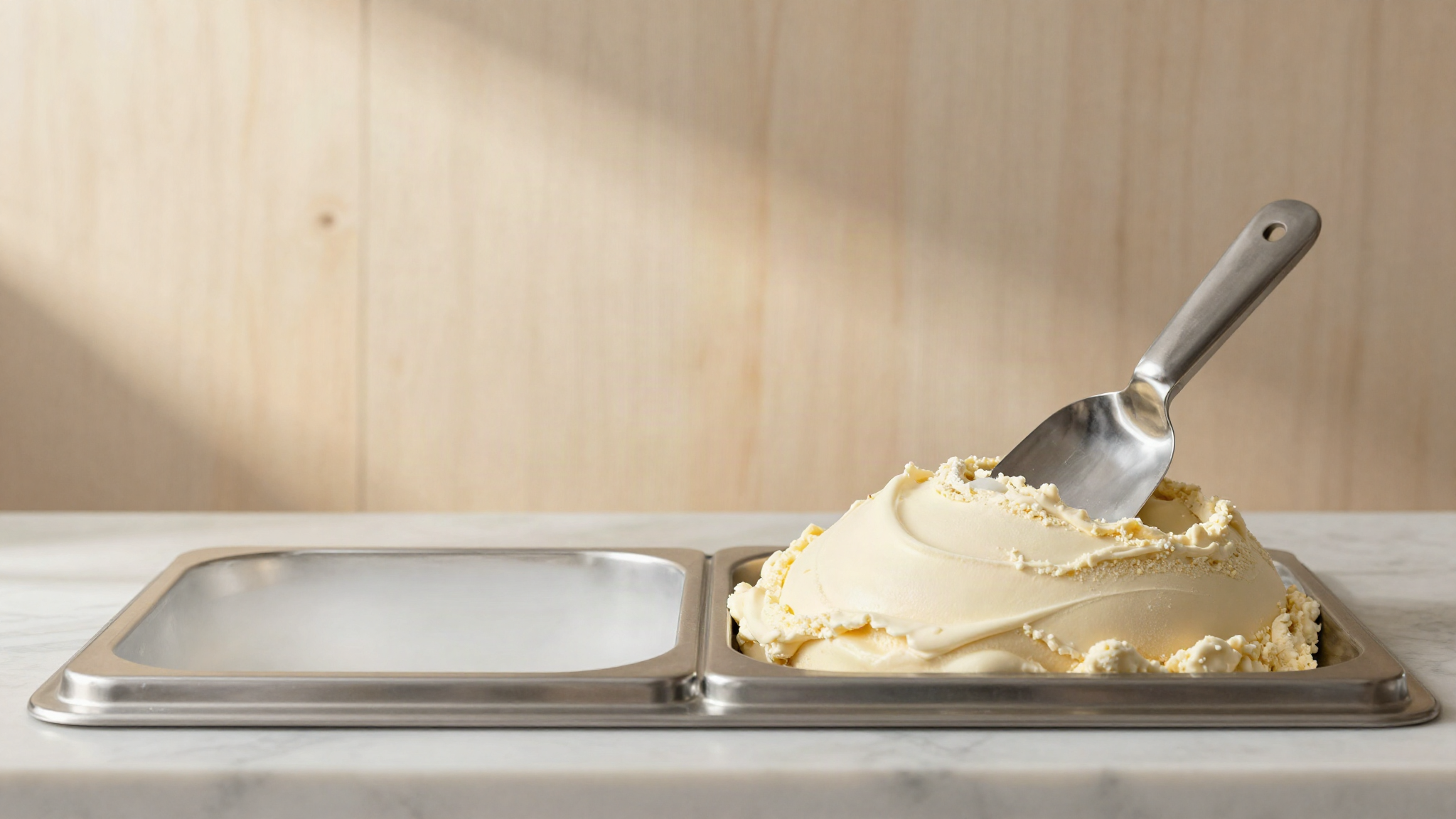 A finished pan of pale gelato in a display case with a stainless steel spade resting on top