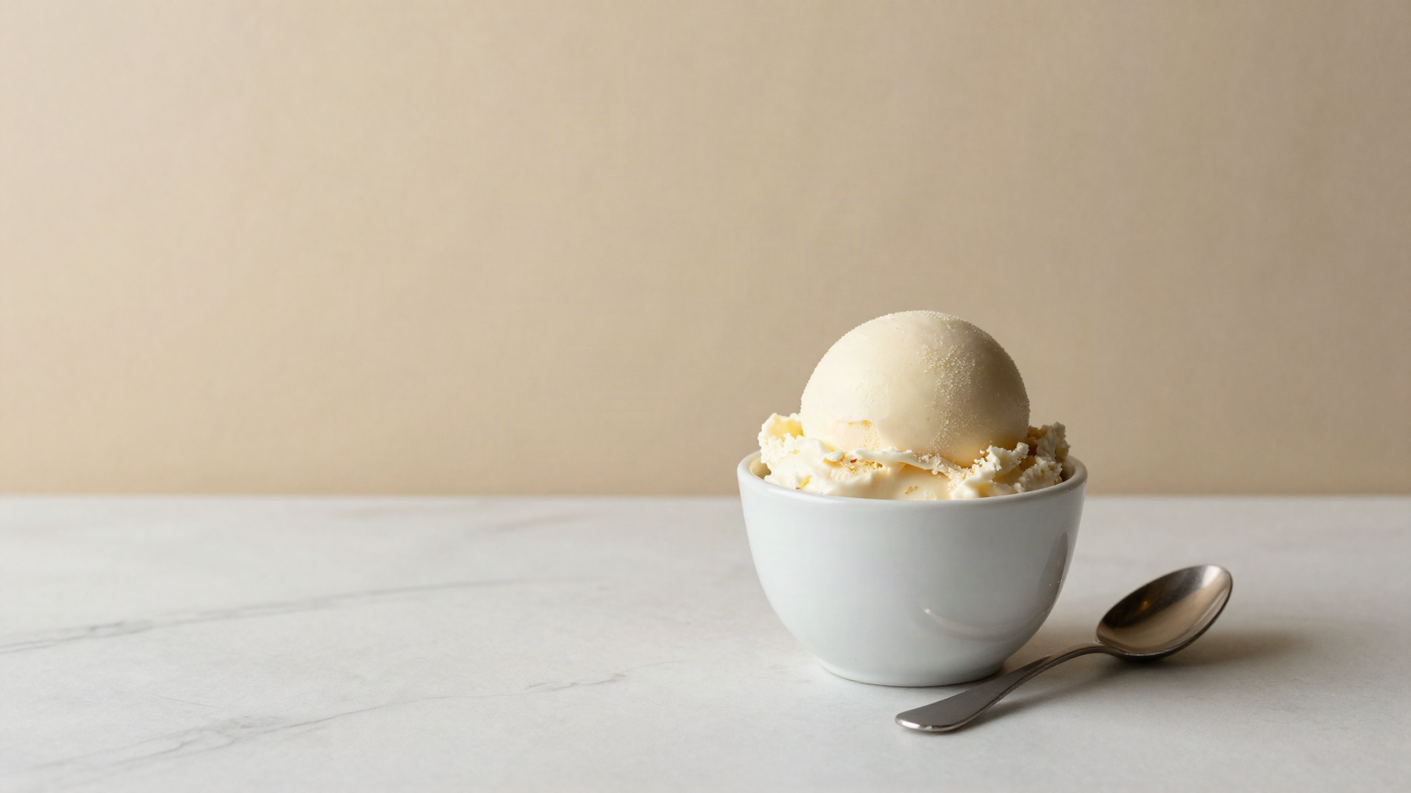 A dense scoop of pale ivory gelato in a small white ceramic cup with a metal spoon on marble