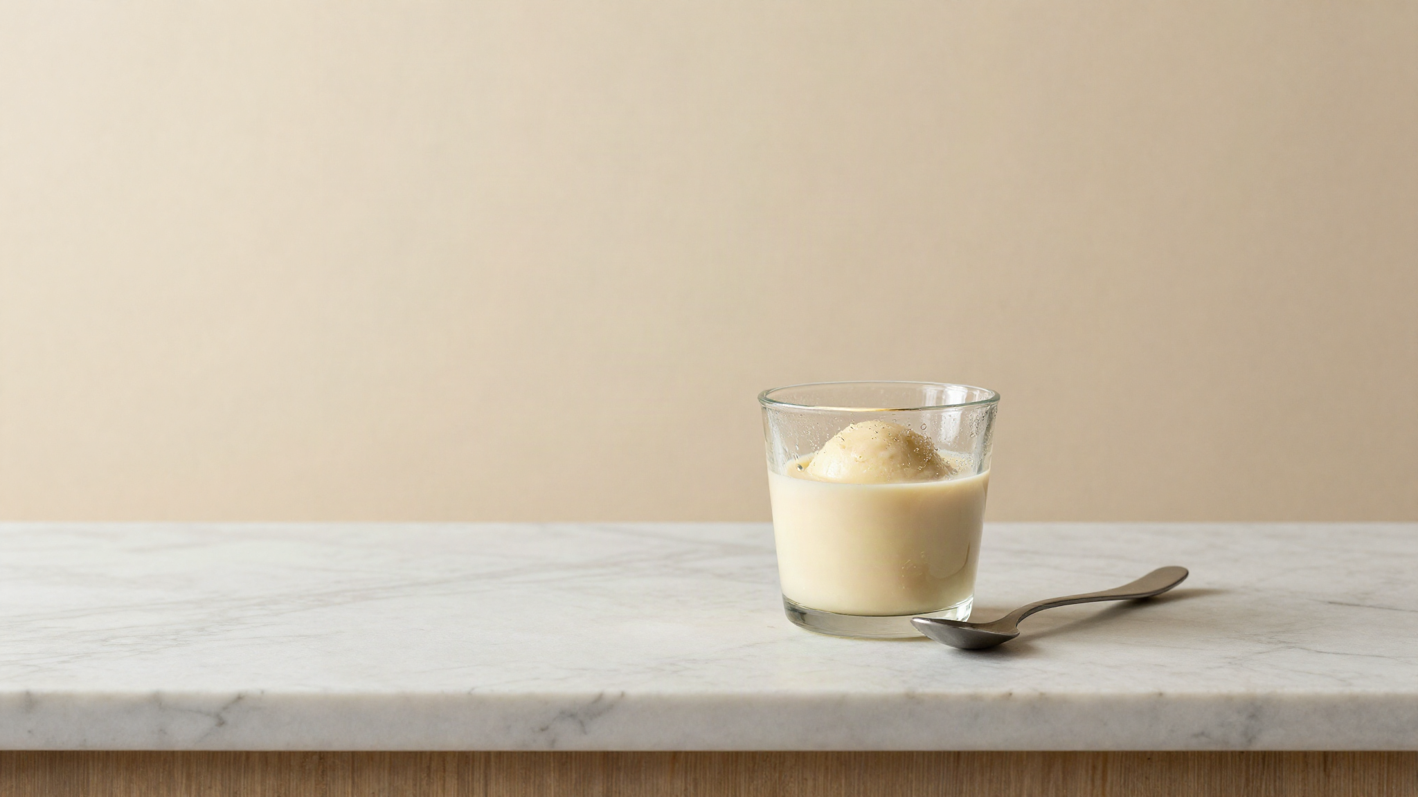 A small glass beaker of pale liquid gelato mix with condensation on the glass and a stainless tasting spoon beside it
