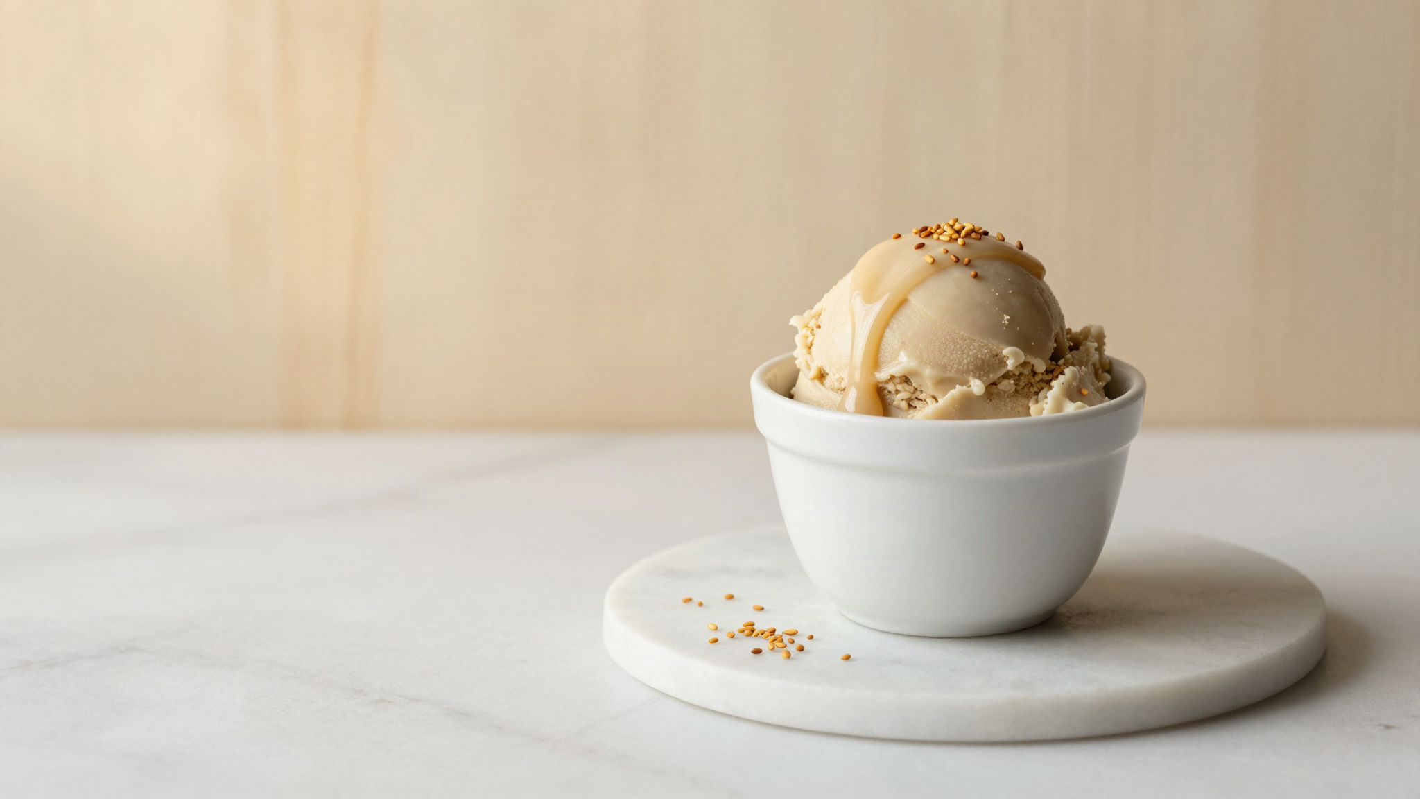 A small white ceramic cup of pale sesame gelato finished with a drizzle of tahini and toasted sesame seeds