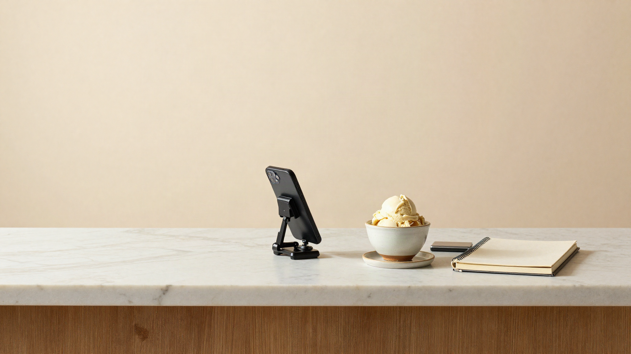 Smartphone on a small tripod on a marble counter framing a ceramic cup of gelato beside a notebook and a small scale