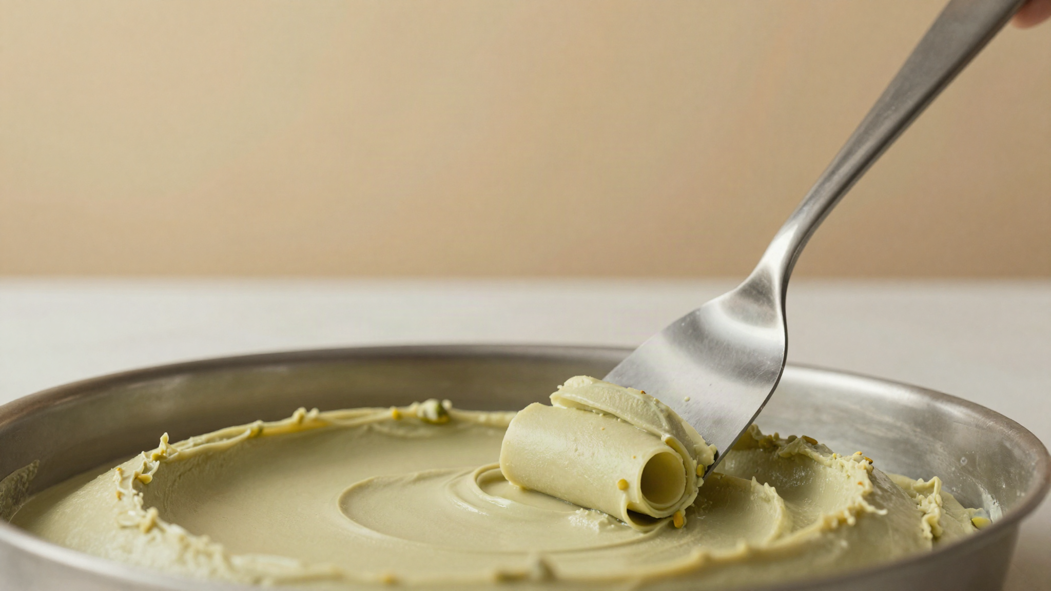 Stainless steel spatula drawing a clean ribbon curl across the surface of pale pistachio gelato in a metal pan