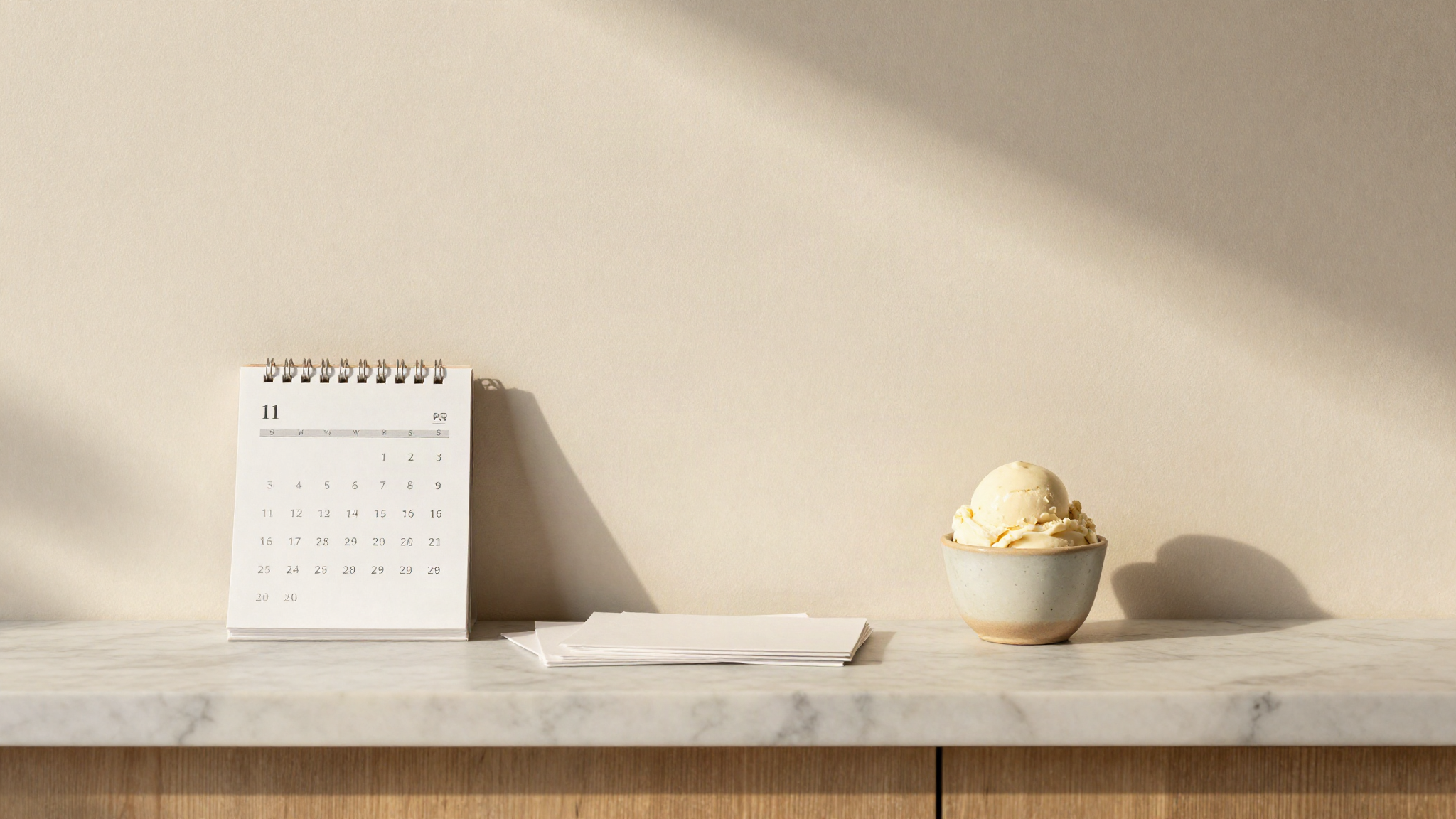 Paper calendar page and a stack of blank index cards on a marble gelateria counter beside a ceramic cup of gelato