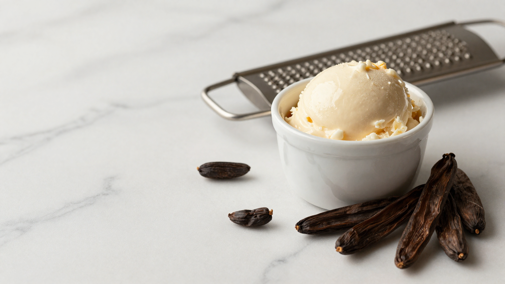 Whole tonka beans on white marble beside a cup of cream-colored gelato and a grater