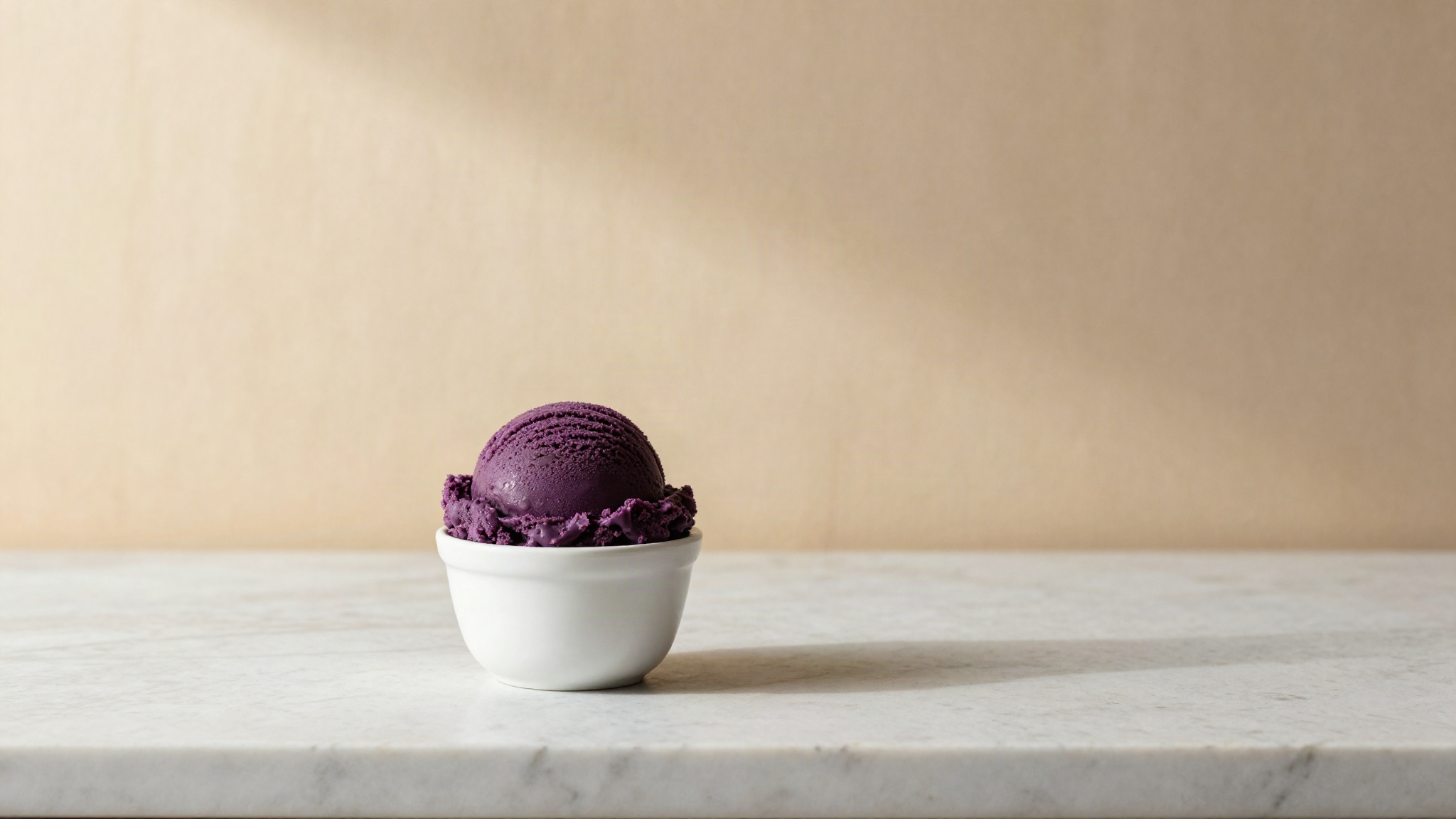 A scoop of deep violet purple gelato in a small white ceramic cup on a marble counter