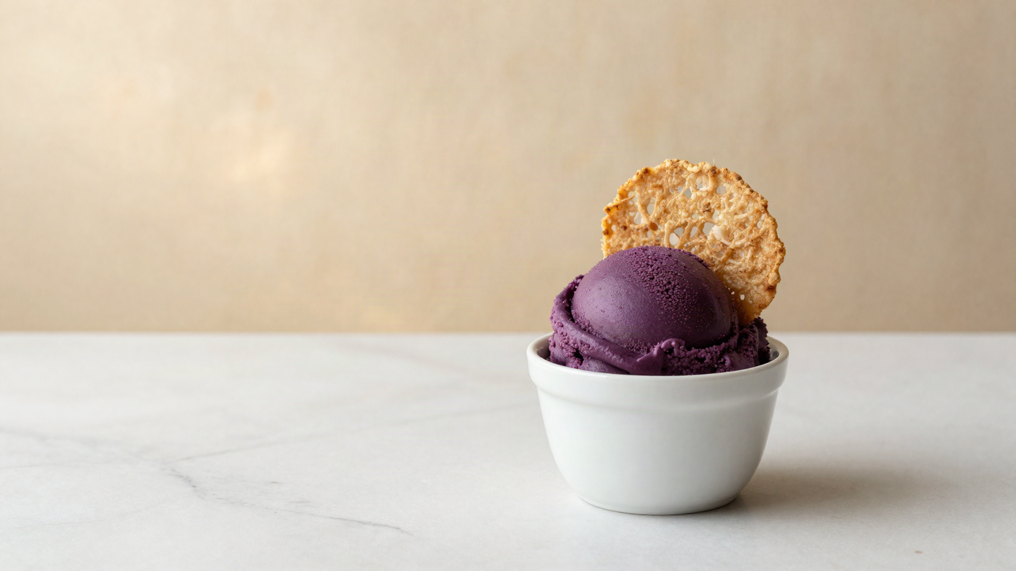 Scoop of deep violet ube gelato in a white ceramic cup with a thin wafer on marble