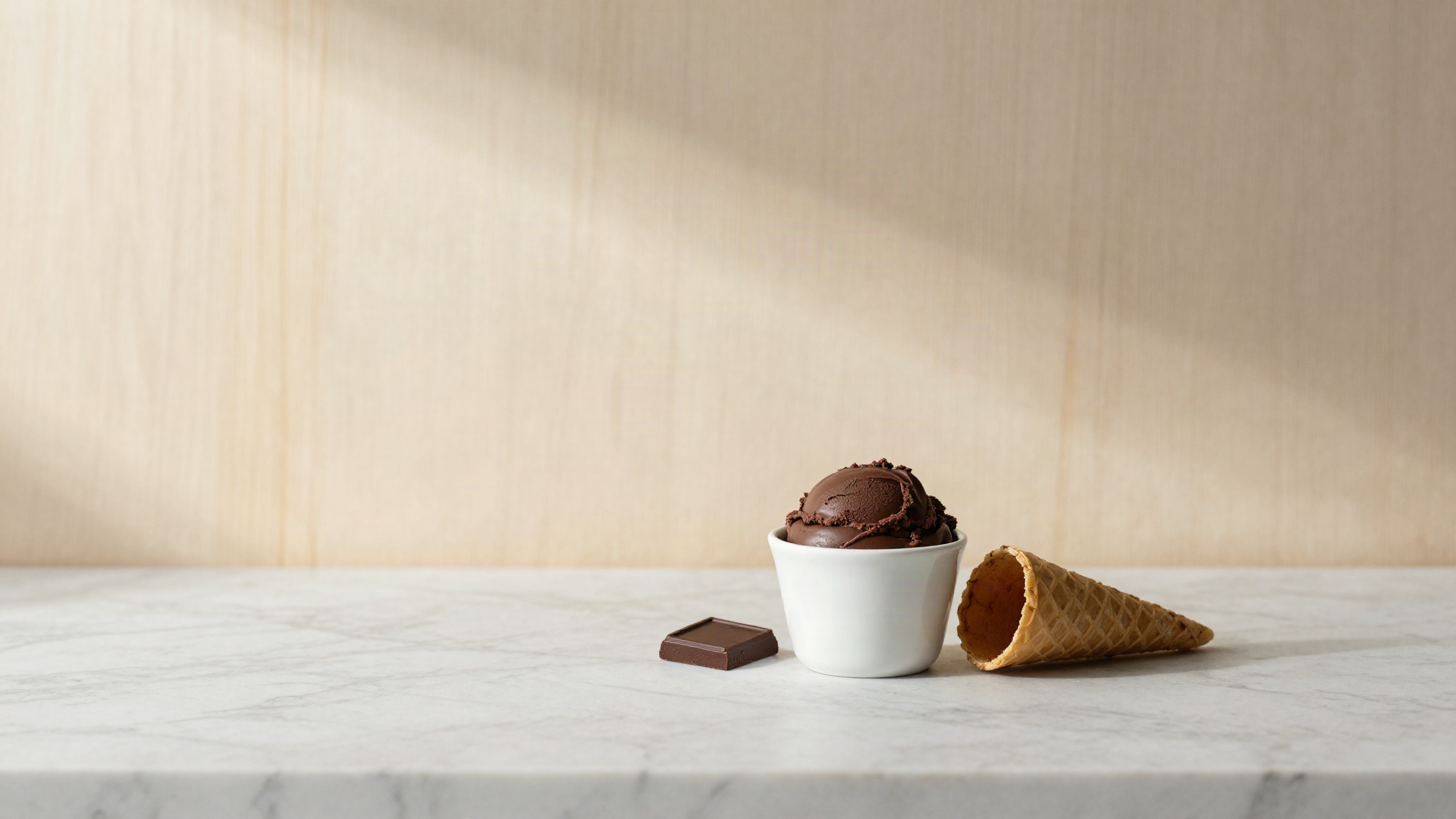 A small white ceramic cup of dark chocolate gelato beside a cone, both on a marble counter, with a square of dark chocolate in soft warm light