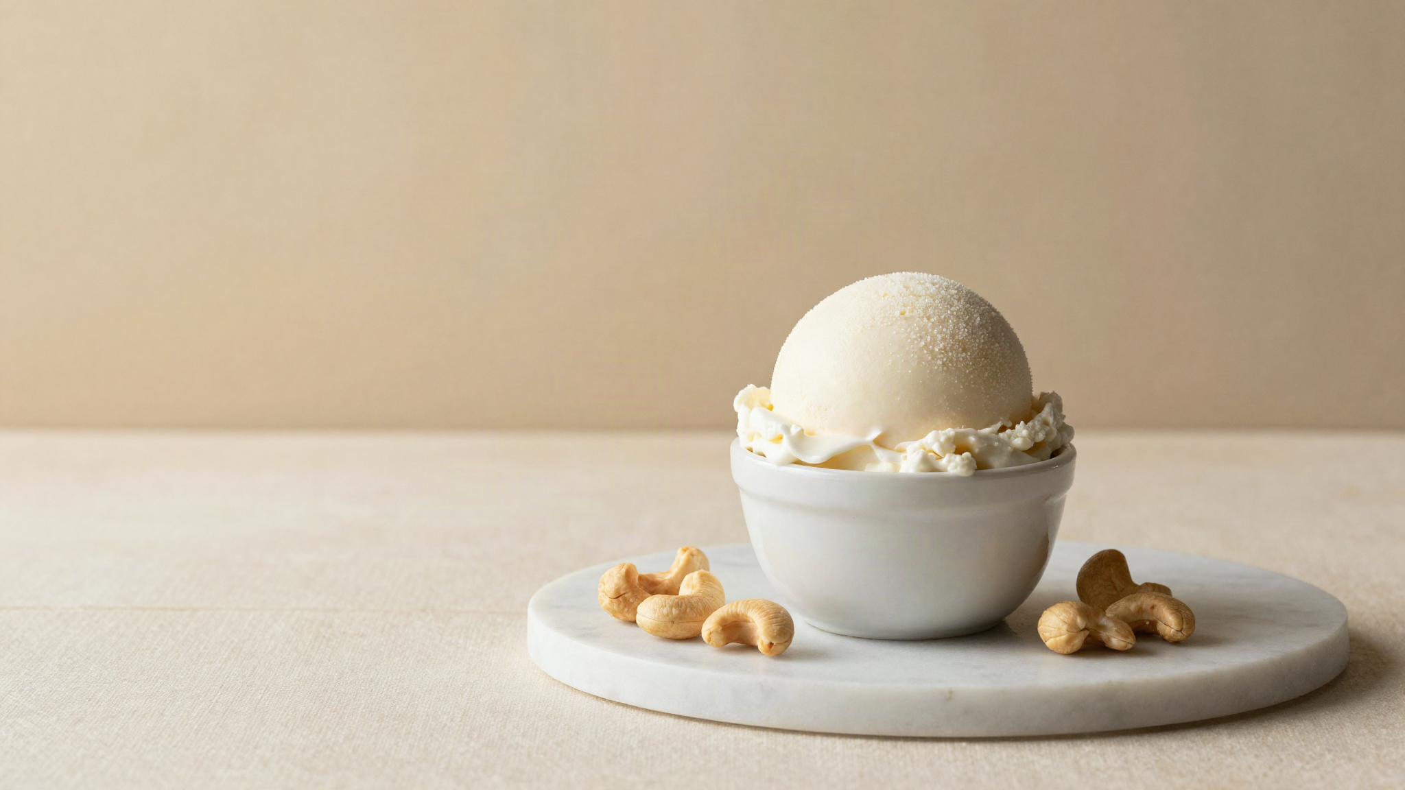 A clean rounded scoop of white vegan gelato in a small white ceramic cup on marble with a few whole cashews beside it