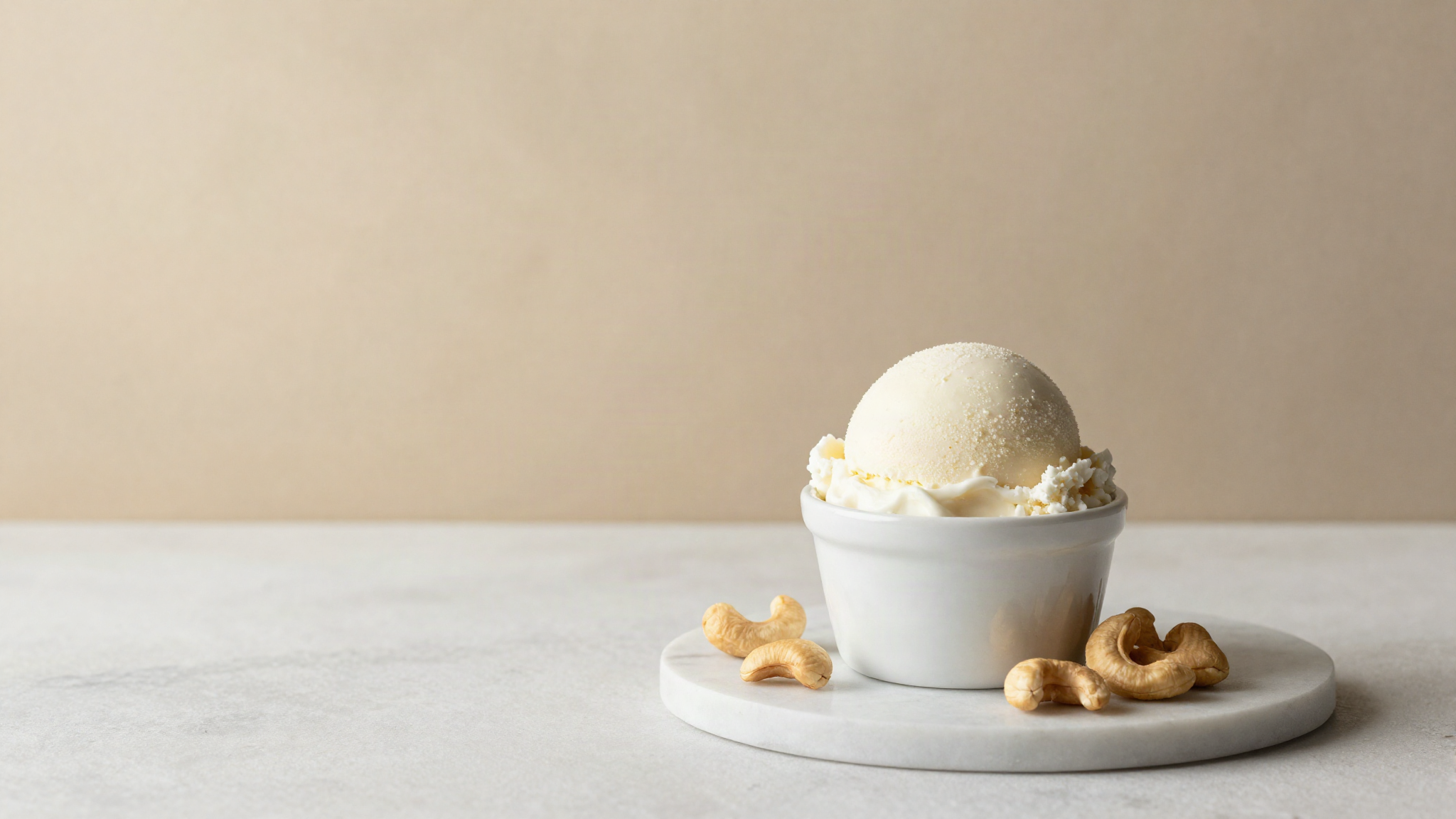 A clean rounded scoop of white vegan gelato in a small ceramic cup on marble, with a few whole cashews beside it
