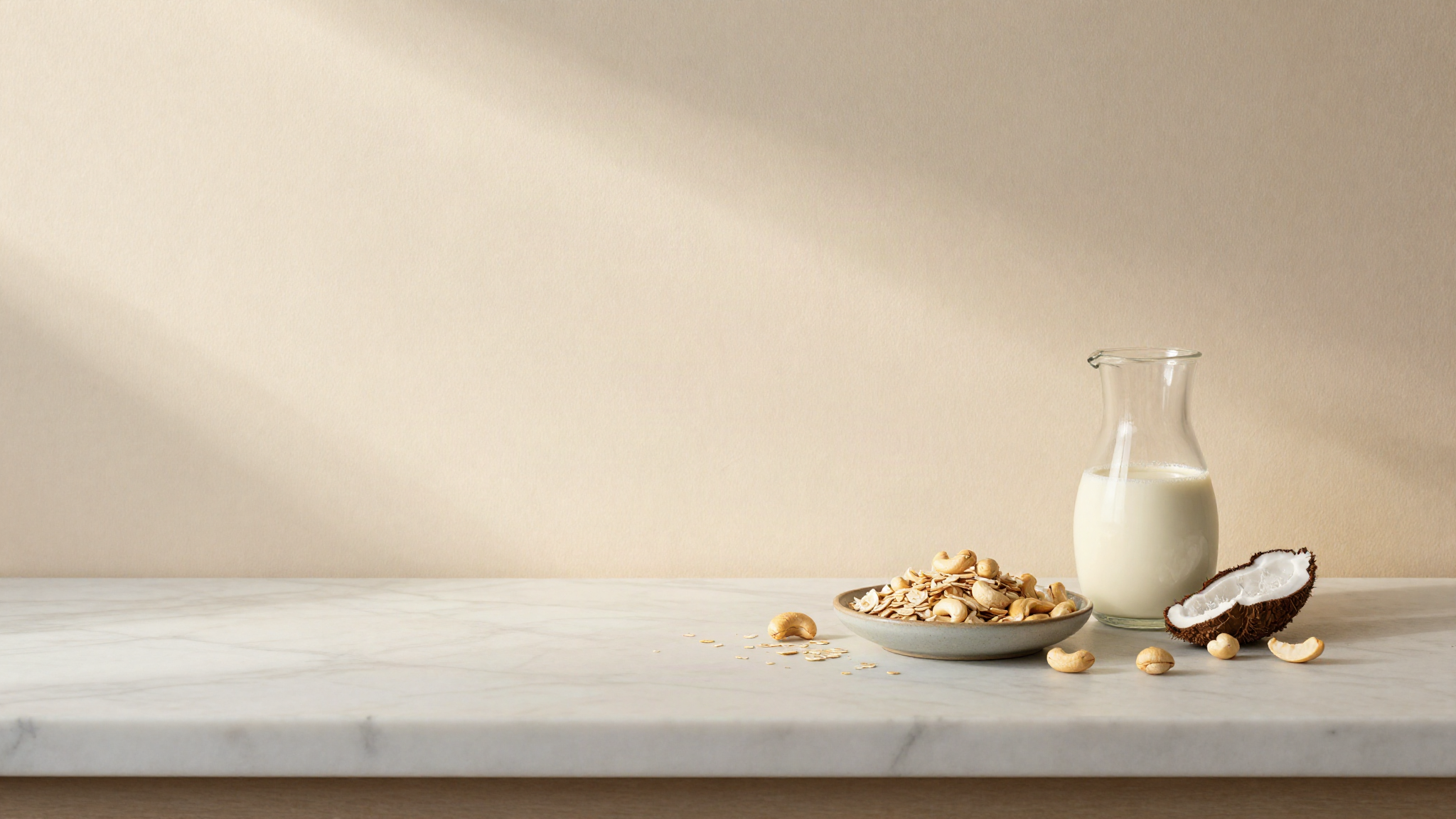 Small ceramic dishes of rolled oats, whole cashews and grated coconut beside a jug of oat milk on a marble counter