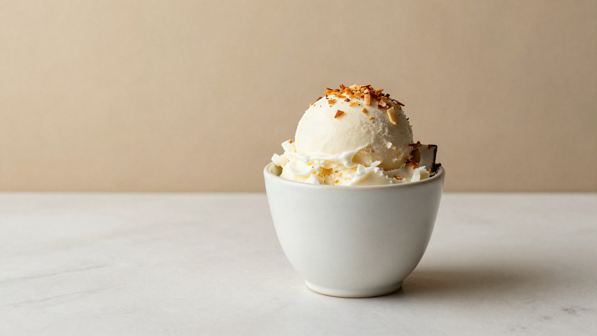 Creamy vegan coconut gelato in a refined ceramic cup topped with toasted coconut flakes on marble