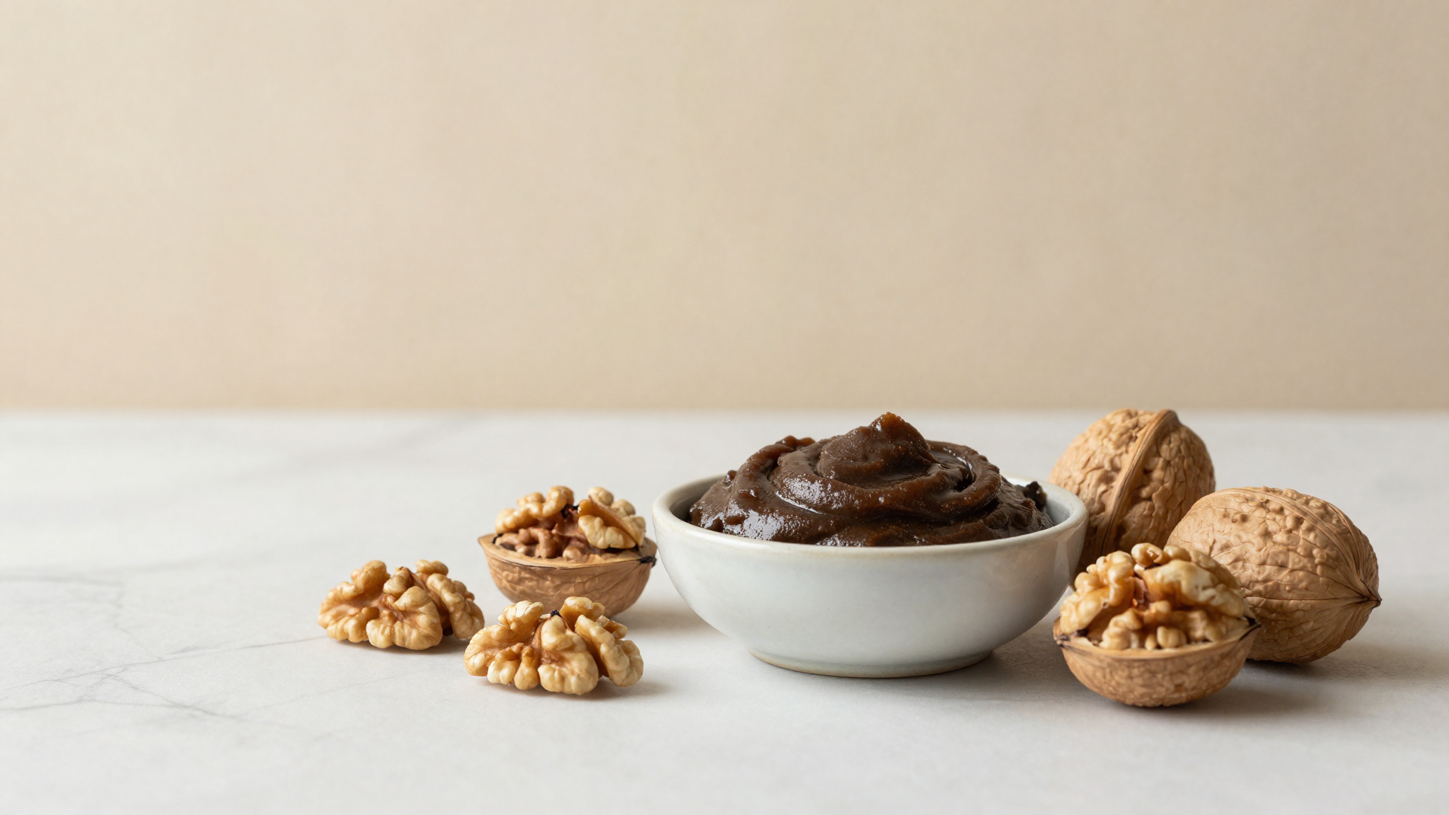 Shelled walnuts and a small ceramic bowl of smooth dark walnut paste on a marble surface