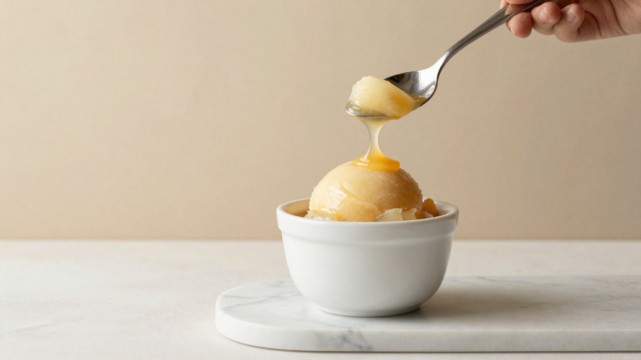 A glossy smooth fruit sorbet quenelle in a white ceramic cup on marble, spoon lifting a silky ribbon