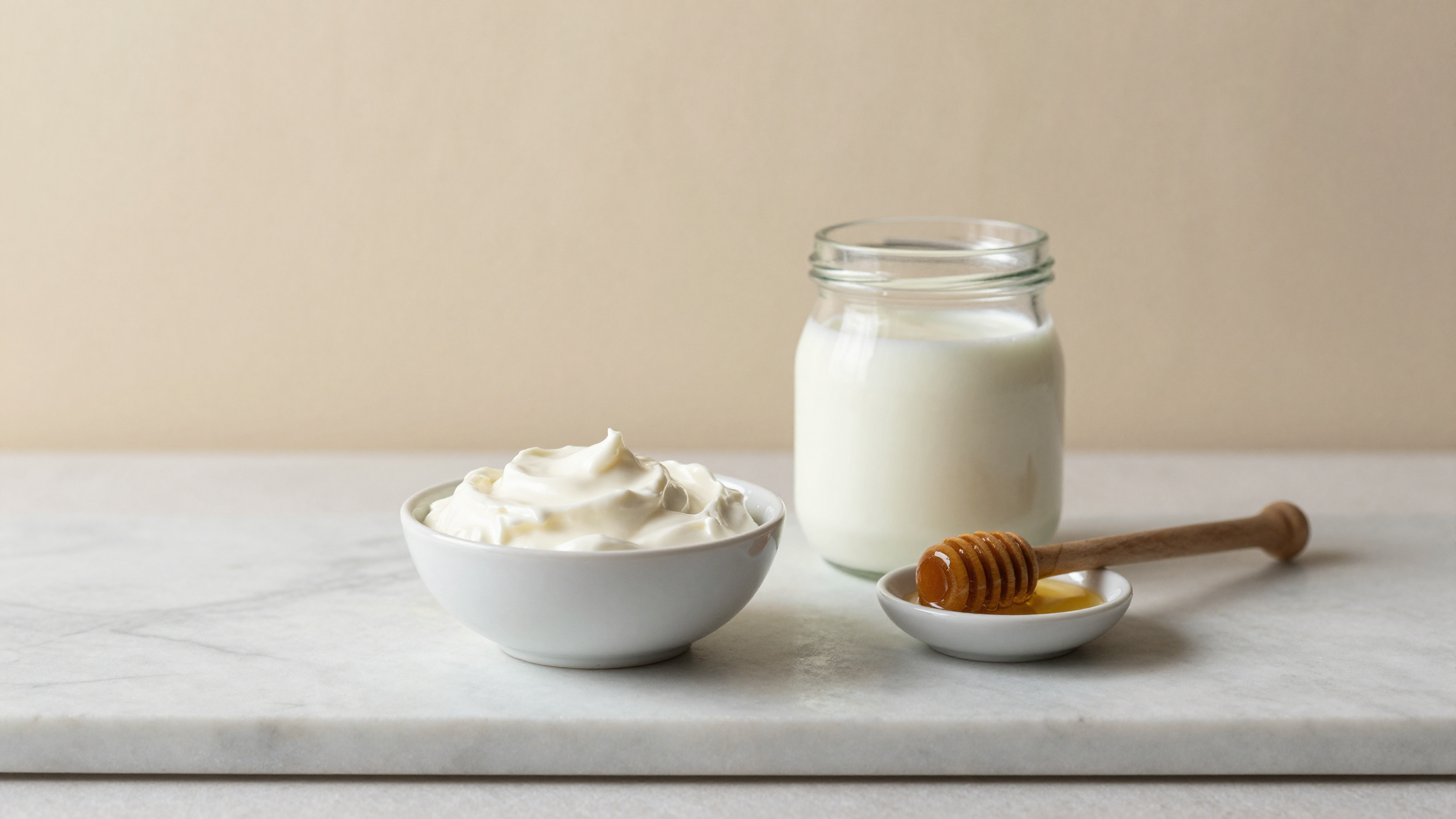 Bowl of strained Greek yogurt beside glass jar of fresh whole milk and small dish of acacia honey on marble counter